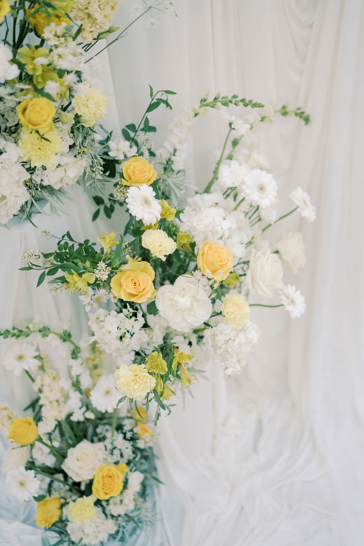 A loose arrangement of yellow roses and white flowers sits against softly draped white fabric.