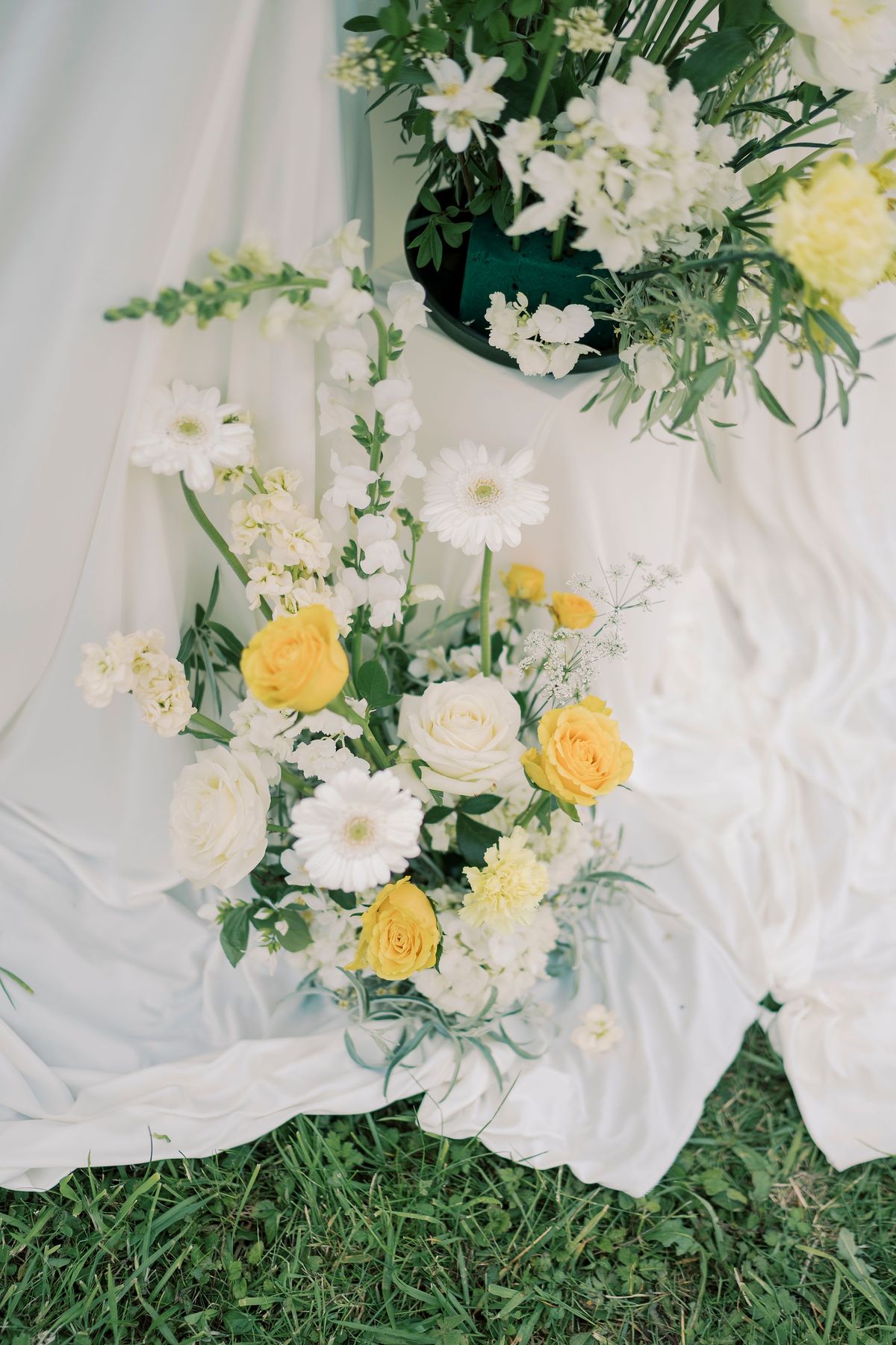 An arrangement of yellow and white flowers with green foliage rests on white draped fabric spread over grass.