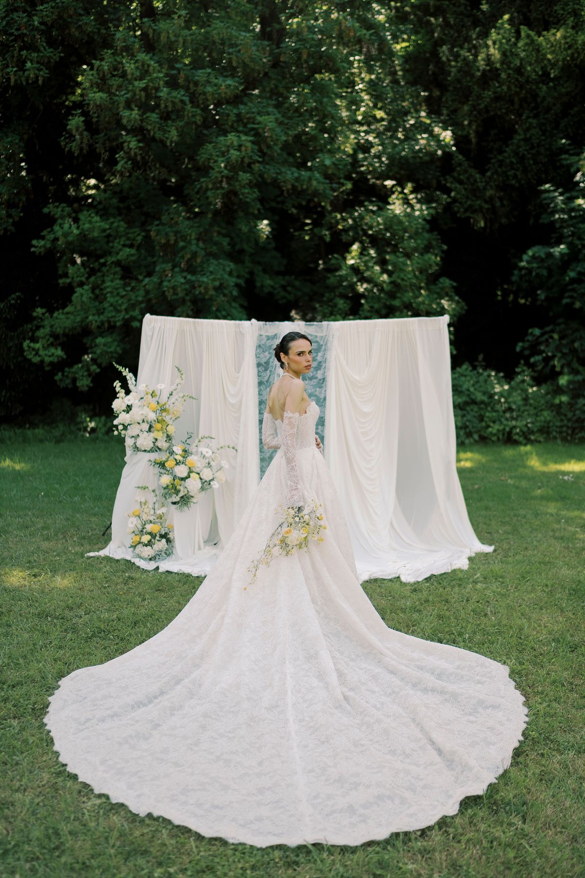 A bride in a lace gown with a very long train stands on a lawn in front of a white draped backdrop, looking back over her shoulder.
