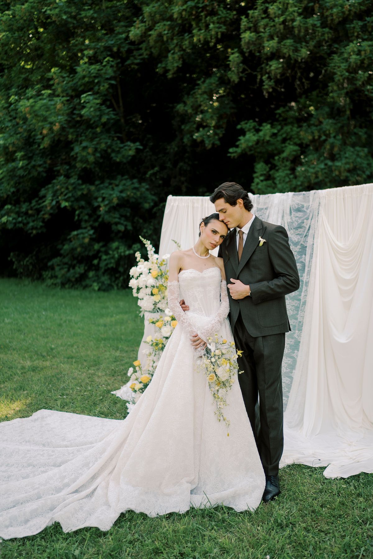 The couple stand close together on grass, the bride holding a bouquet, in front of hanging white drapes and trees.