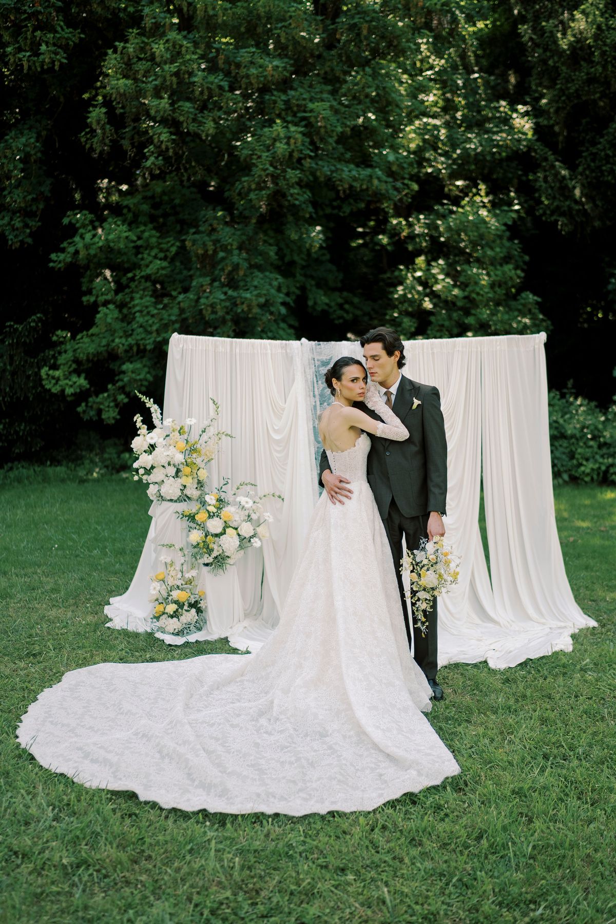 The couple embrace on a lawn in front of white draped fabric and floral arrangements, the bride's long train spread behind her.
