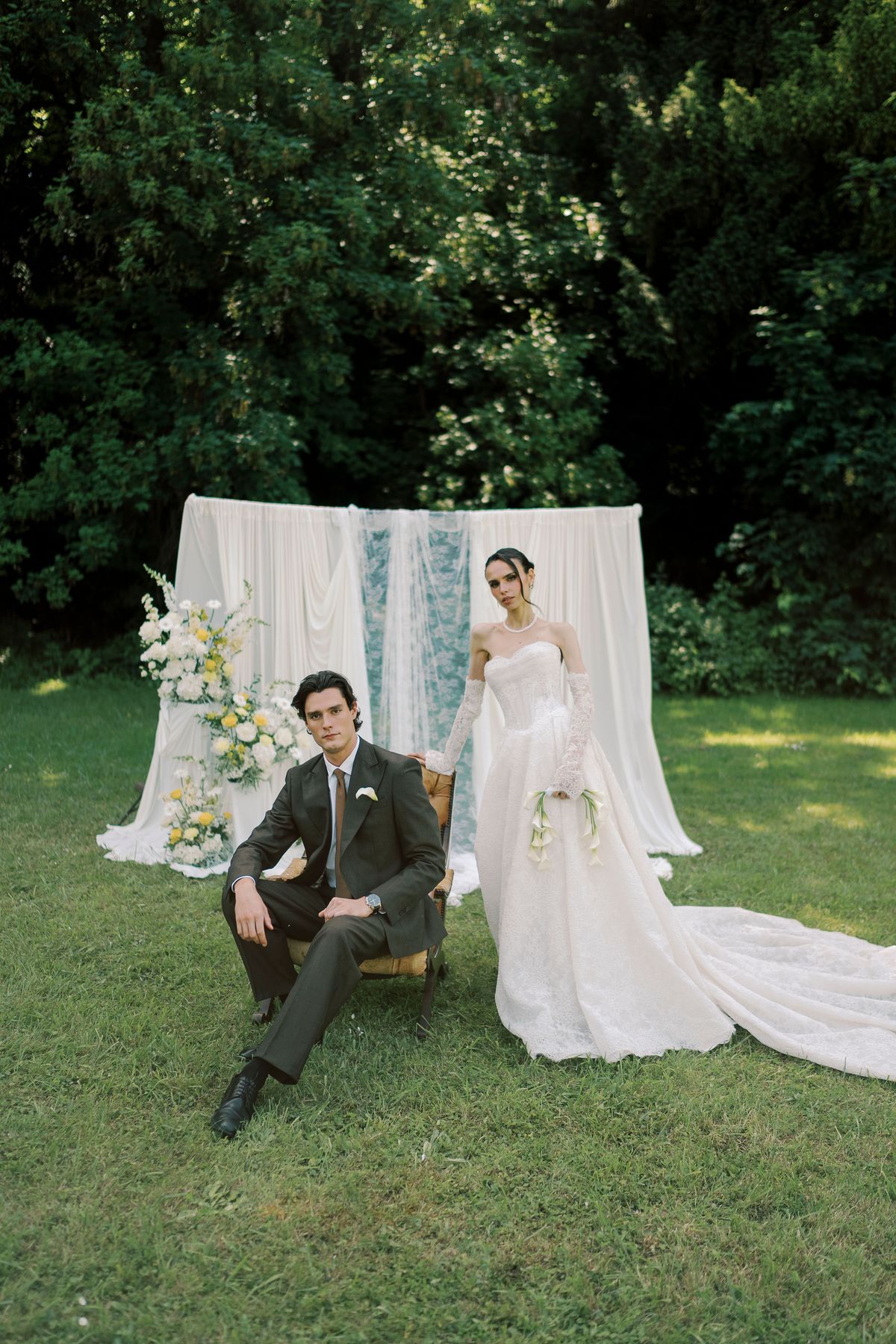 The groom sits on a chair in a dark suit while the bride in a long gown stands beside him on a lawn before draped fabric.