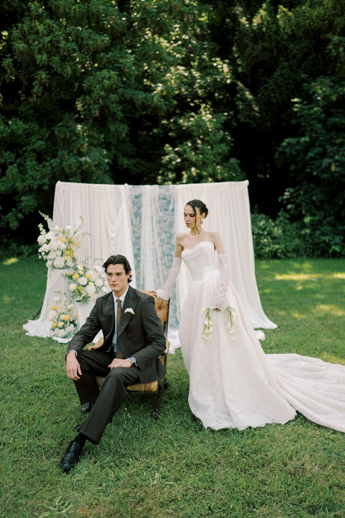 The groom sits in a chair on the lawn while the bride stands beside him holding flowers, in front of hanging white drapes.