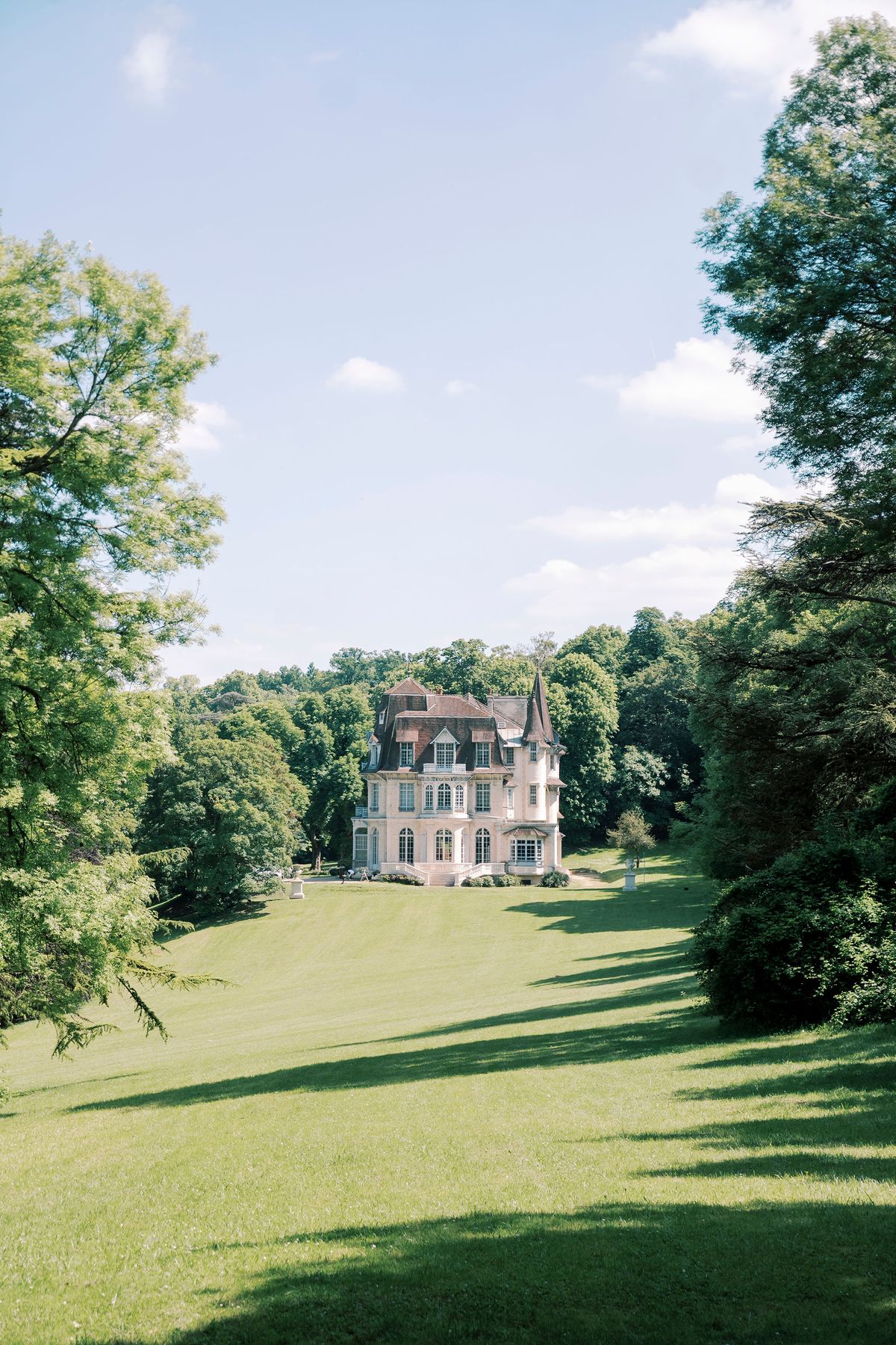 A distant chateau sits among trees at the end of a long green lawn under a blue sky with light cloud.