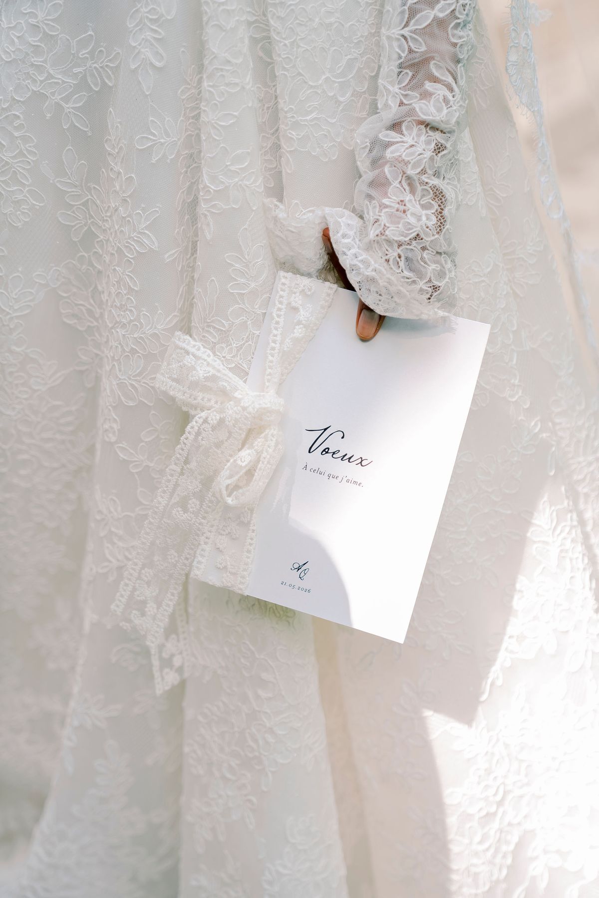 The bride, in a lace gown with sheer lace sleeves, holds a white vows booklet tied with a lace bow, titled "Voeux".