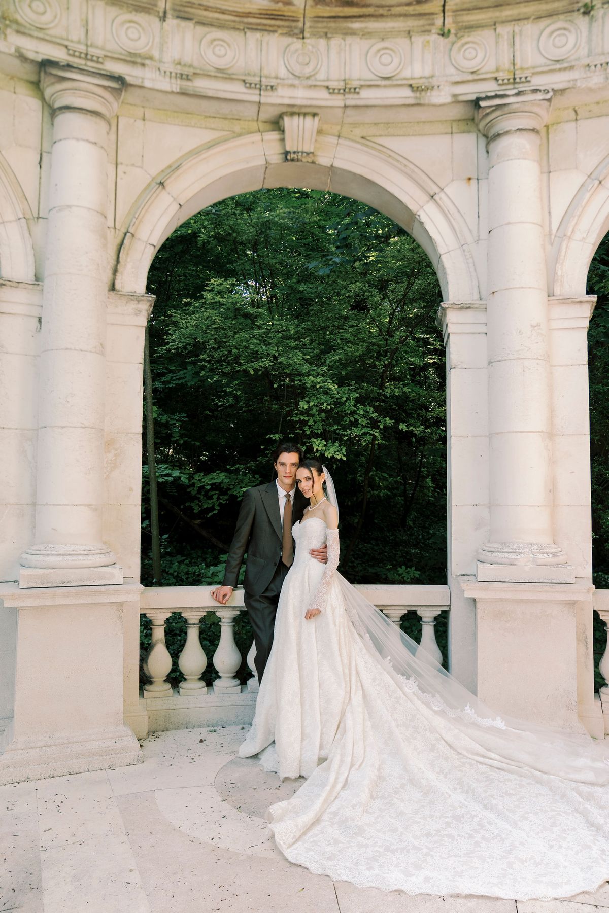 The couple kiss in a stone archway with a balustrade, the bride's very long train spread across the floor.