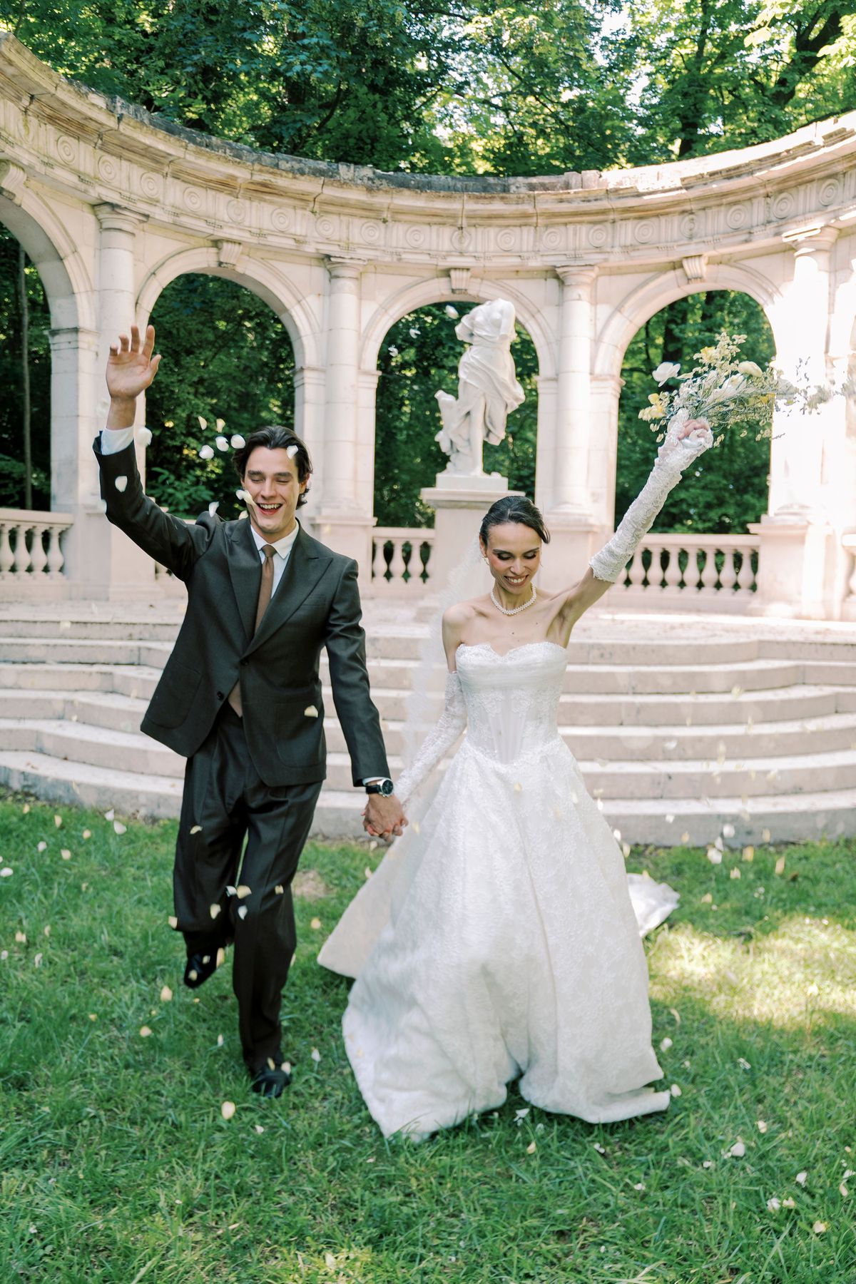 The bride and groom walk hand in hand across the lawn, each with an arm raised and petals falling, in front of a curved stone colonnade with a statue at its centre.