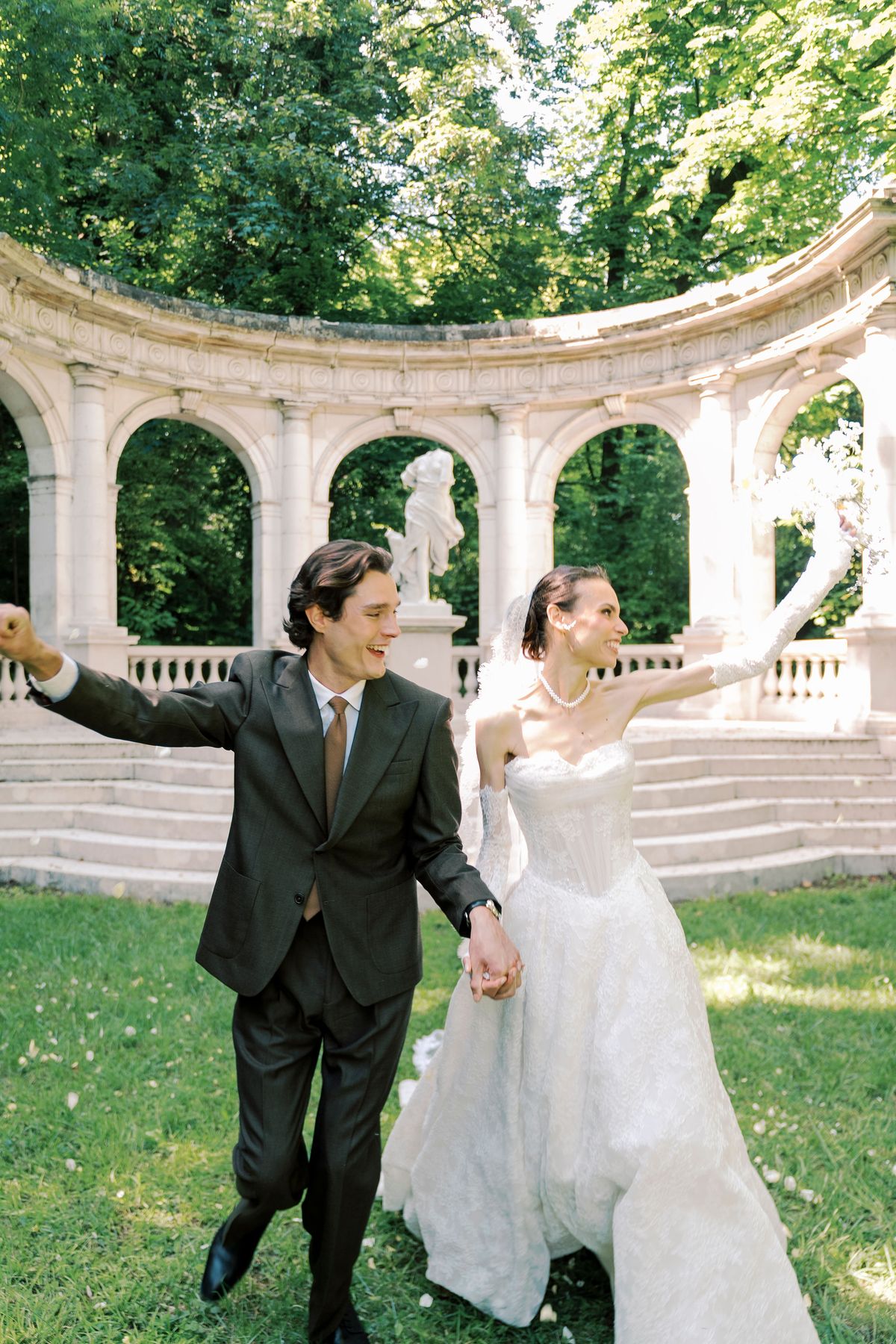 The couple run hand in hand across a lawn, laughing with arms outstretched, in front of a stone colonnade and trees.