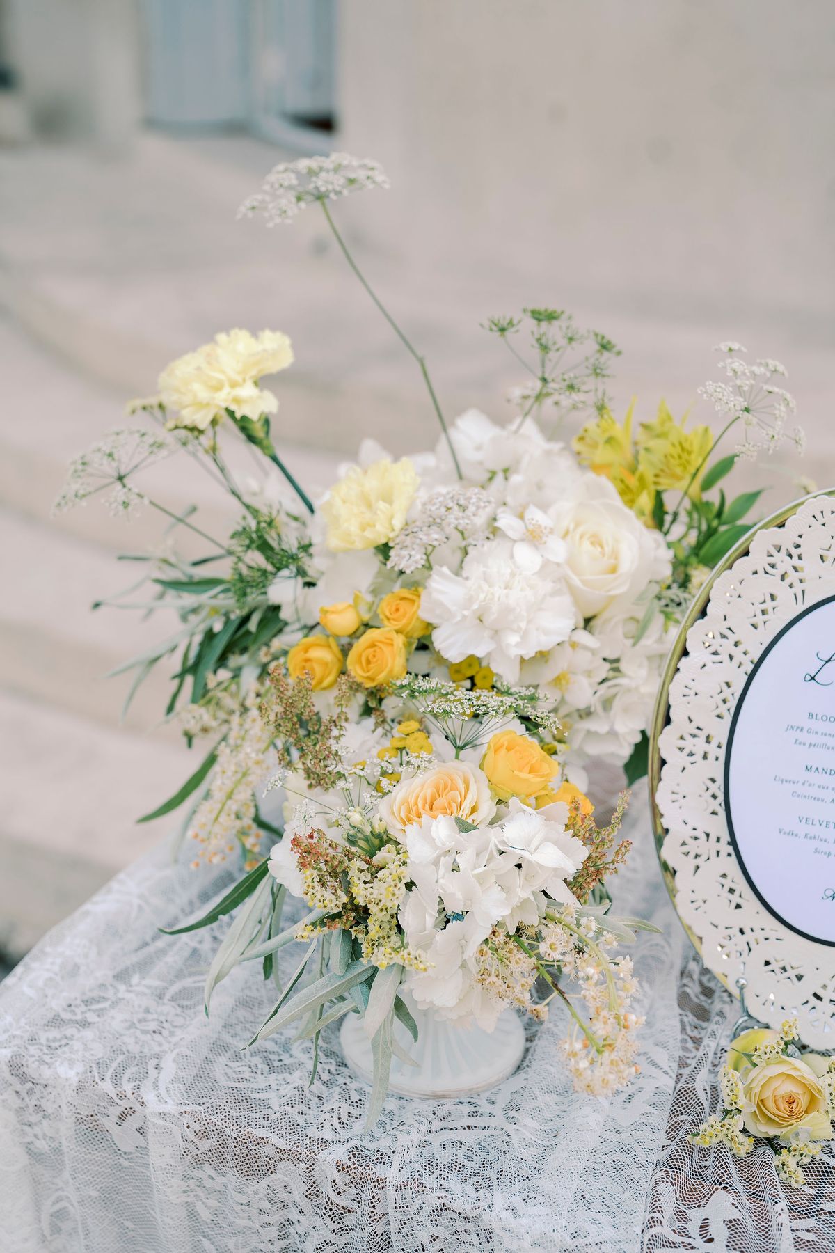A centrepiece of yellow and white flowers and greenery sits on a lace tablecloth beside a lace-edged oval sign.