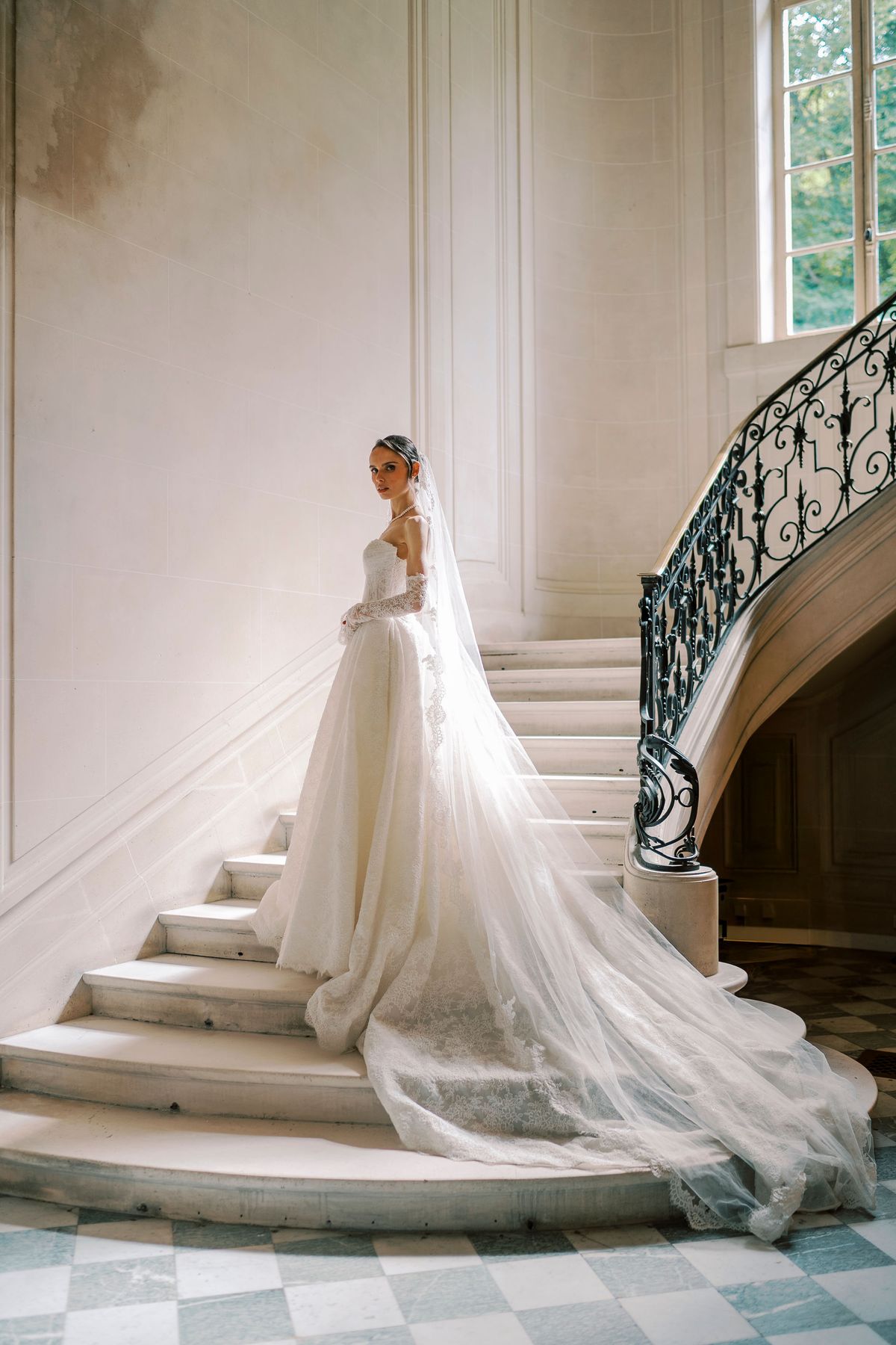 The bride in a white gown with a very long veil stands on a curved stone staircase with an ornate iron railing.