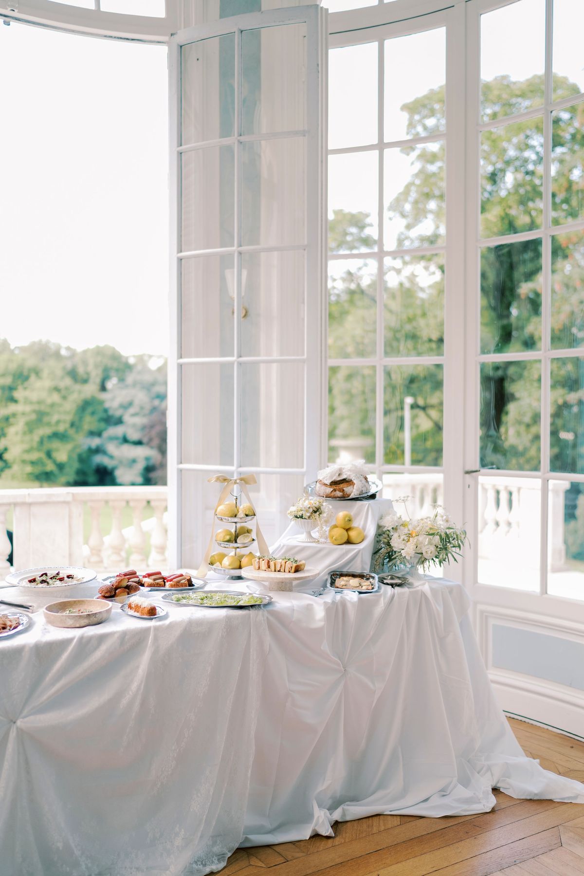 A table covered in a white cloth holds platters of pastries and savoury bites in front of tall glazed windows overlooking trees.