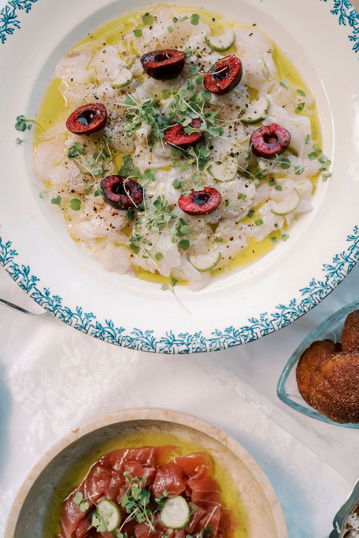 Two plates of sliced raw fish garnished with cherries, herbs and olive oil, one with a blue patterned rim, on a white tablecloth.