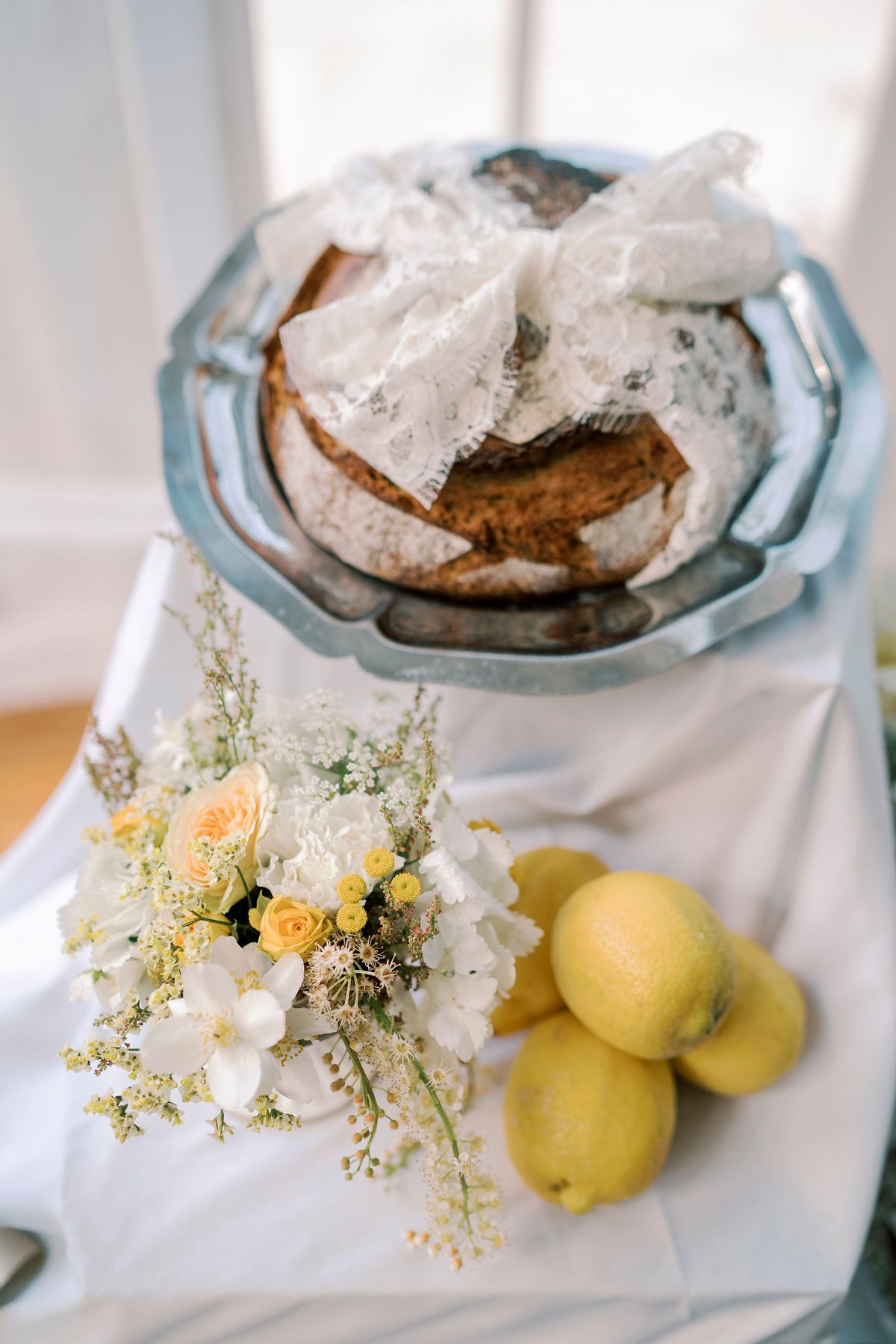 A loaf of bread tied with a lace bow sits on a pewter plate above a yellow and white floral arrangement and several lemons.