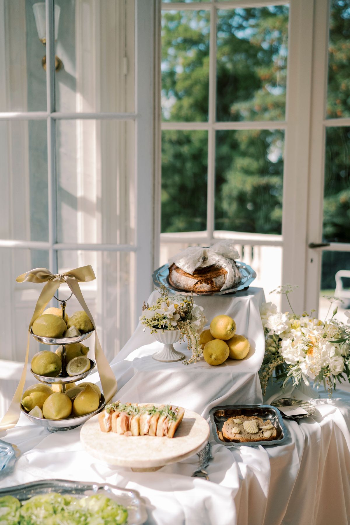 A white-draped table holds tiered stands of pears and lemons, bread, sandwiches and flowers beside tall windows overlooking trees.