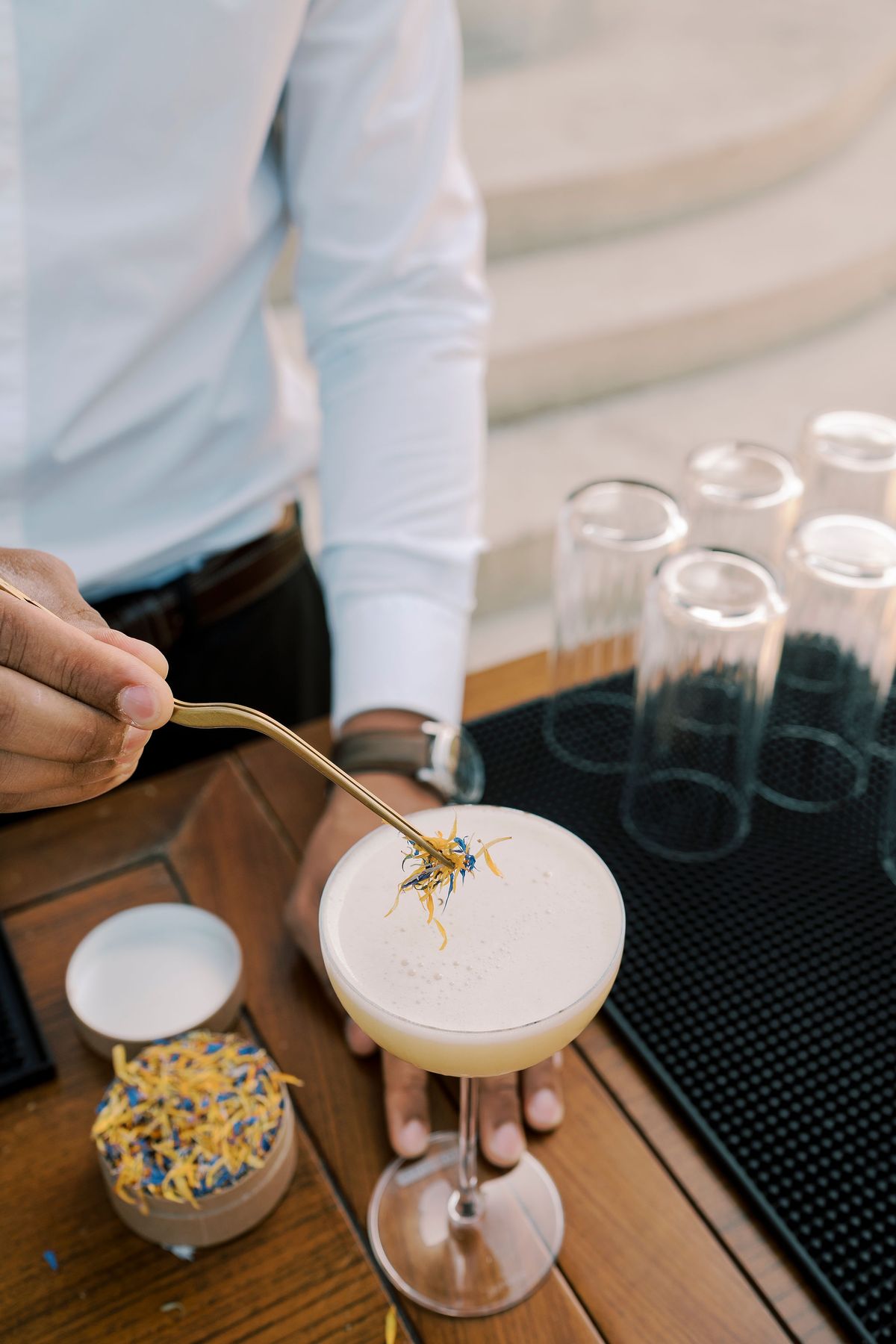 A bartender's hands use a small spoon to garnish a creamy cocktail in a coupe glass, with empty glasses and a dish of dried petals nearby.
