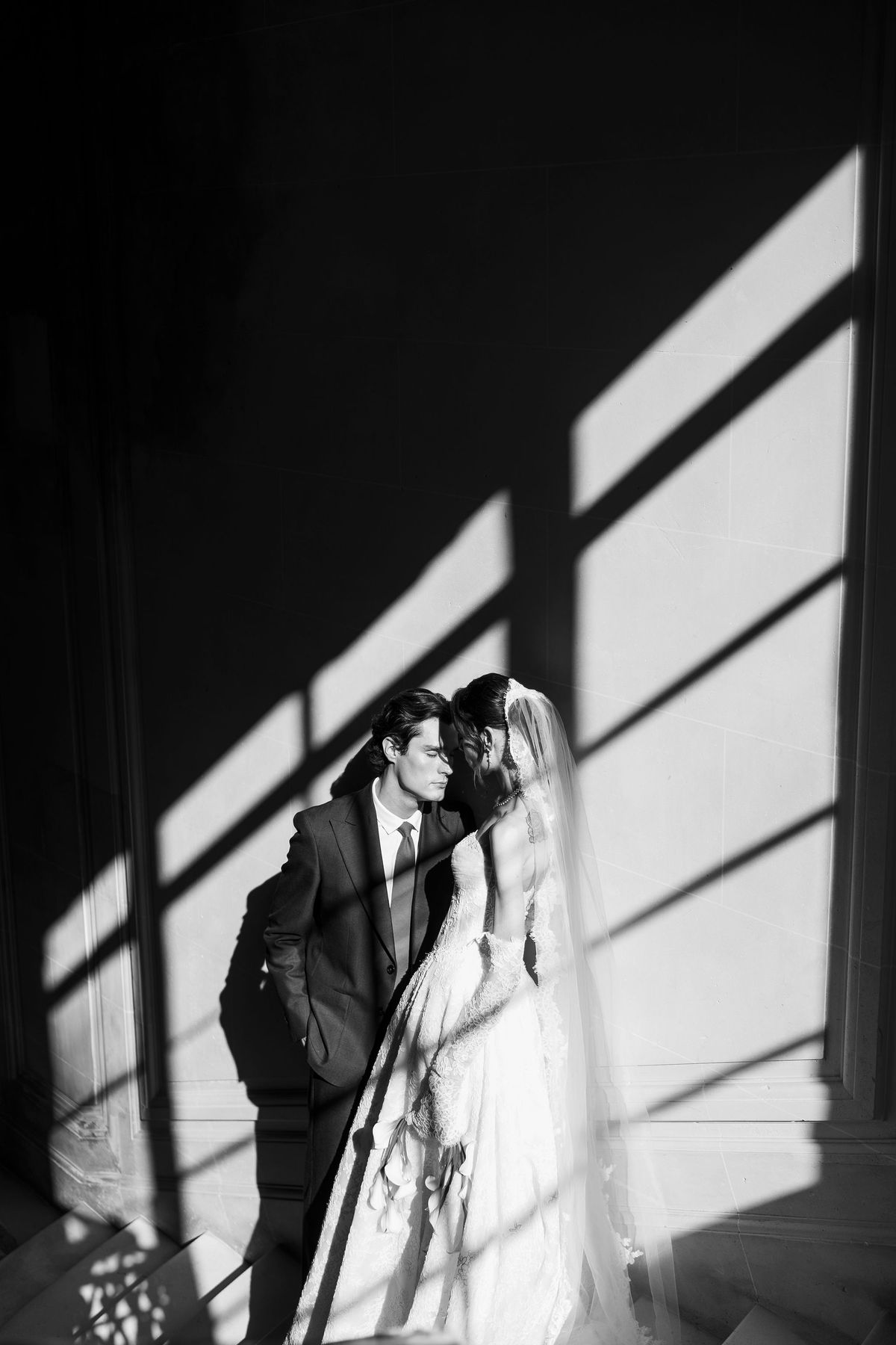 In black and white, the couple embrace and kiss beside a wall patterned with the shadows of tall window frames.