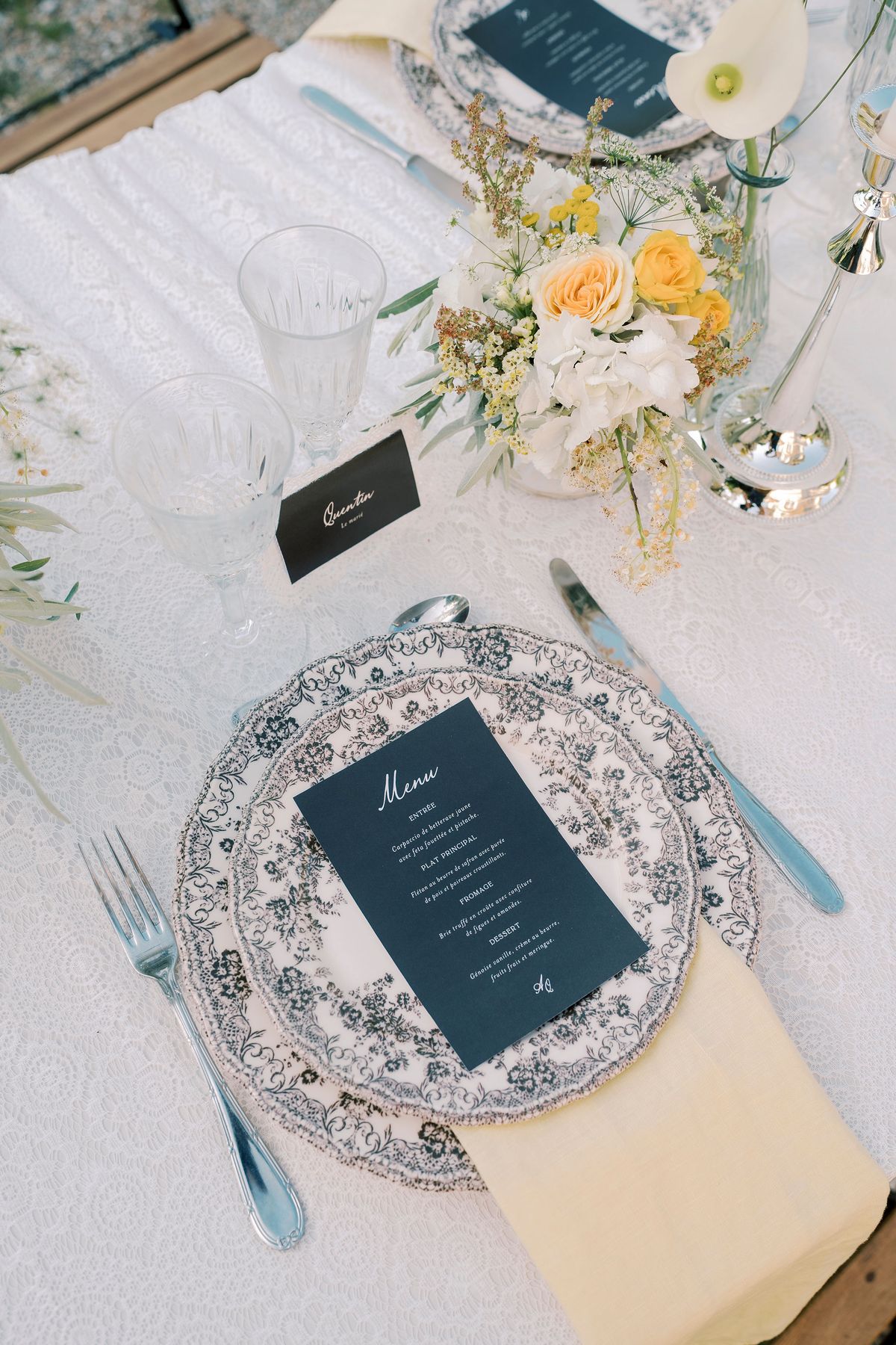 A table setting with a patterned plate, a navy menu card, a yellow napkin, cutlery, glasses and yellow and white flowers.