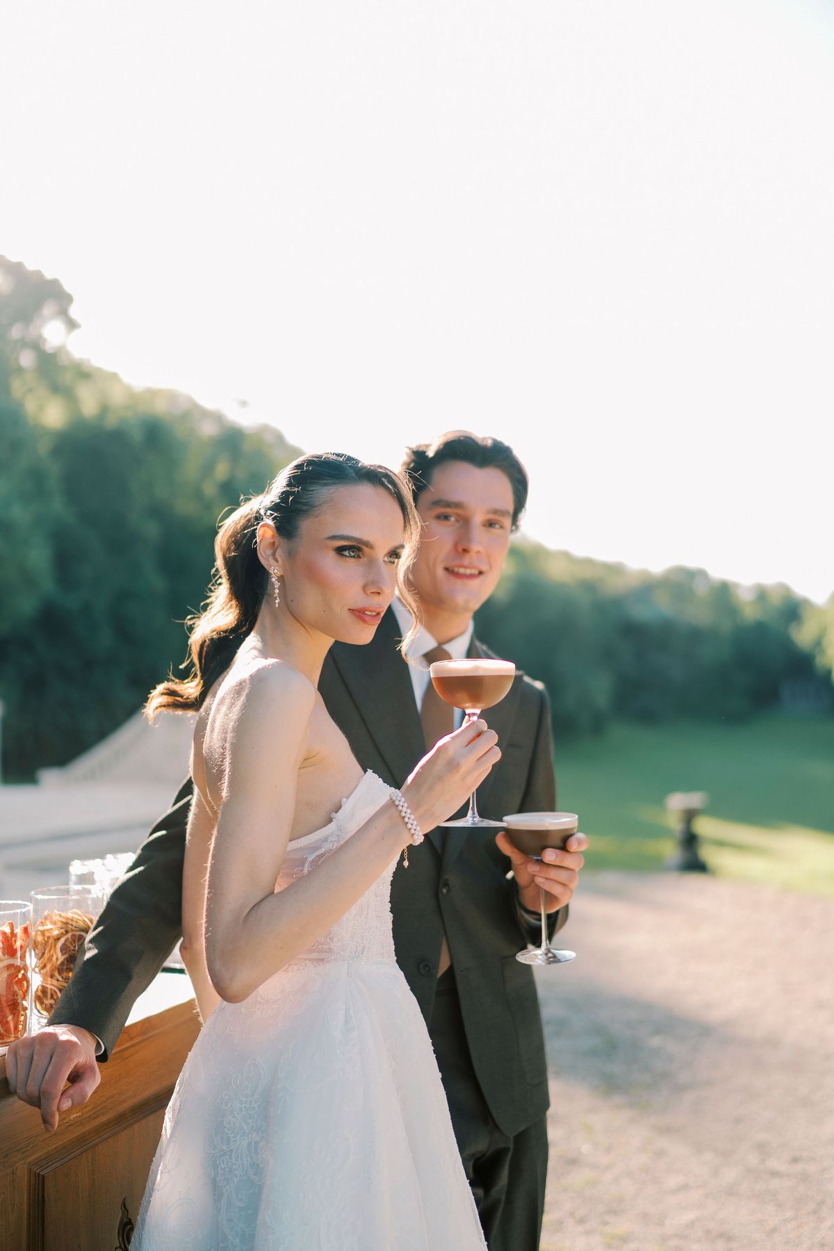 The couple stand close together on a terrace, each holding a cocktail glass, with trees and a pale sky behind them.