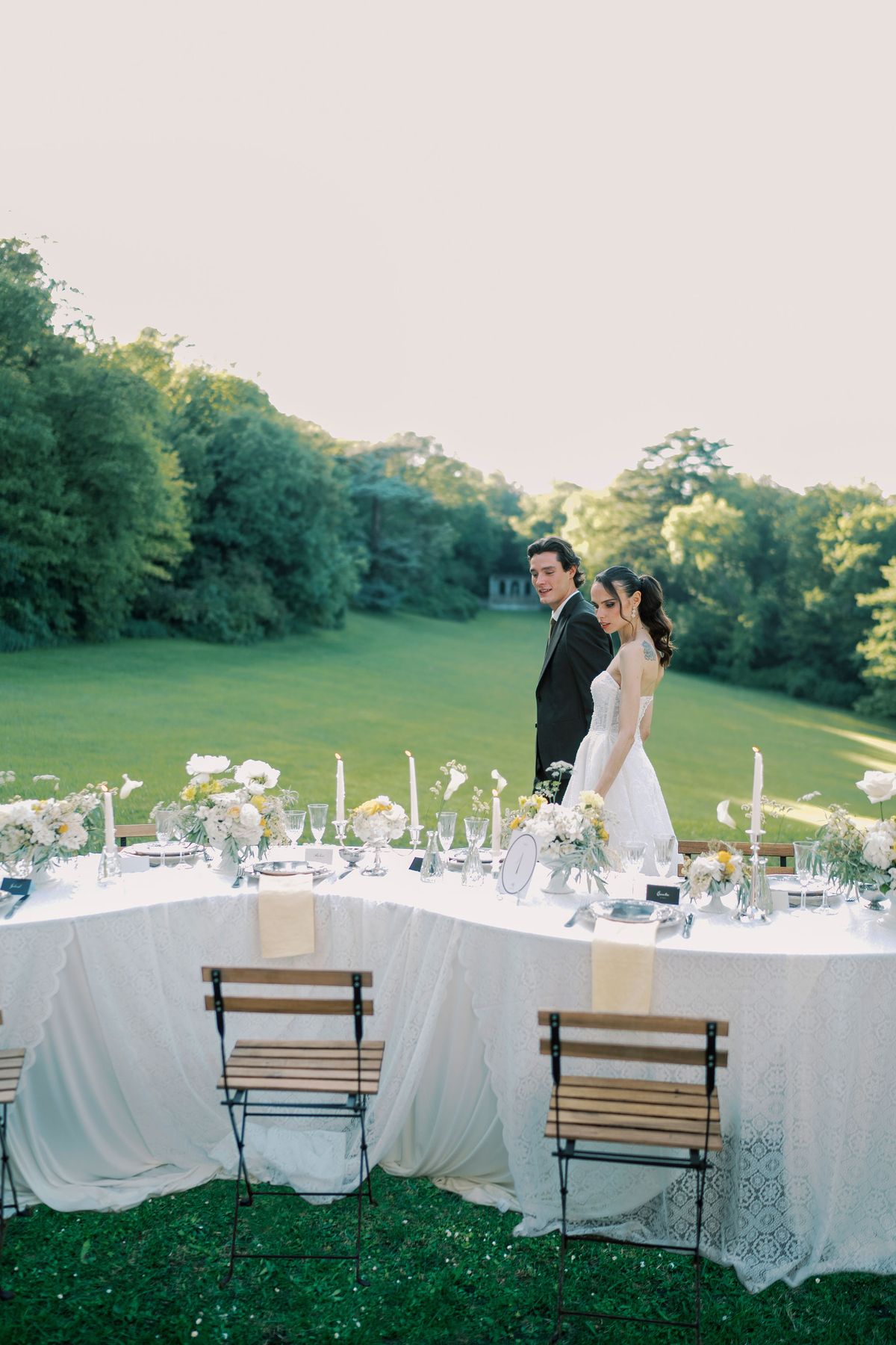 The couple stand together at the far end of a long, white-clothed table set with candles and flowers, on a lawn with trees behind.