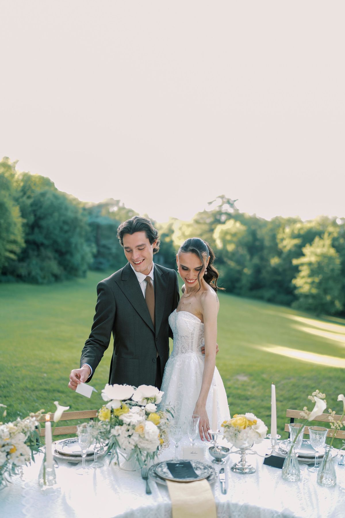 The couple stand at a set table with flowers and candles on a grassy lawn, with wooded hills in the distance.