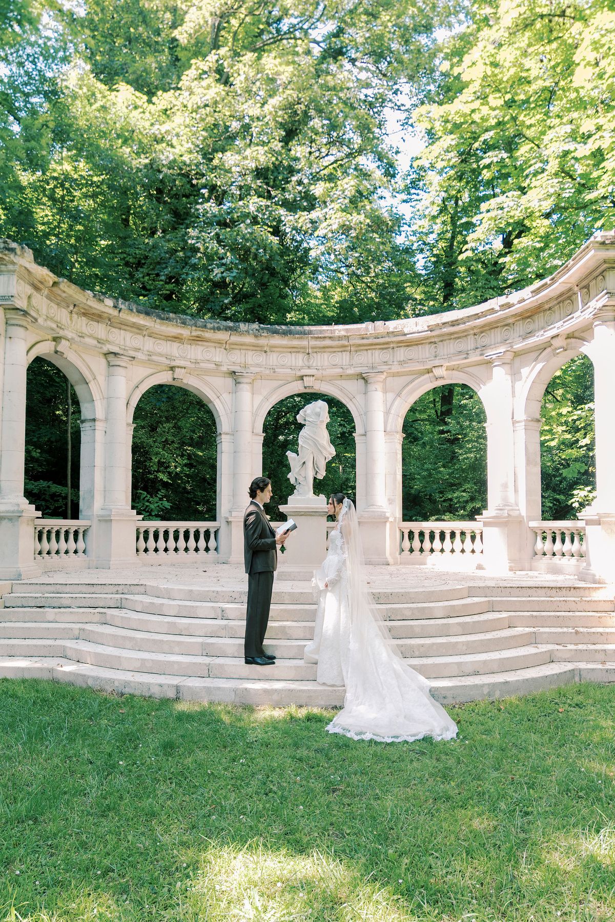 In a wide view, the couple stand together on steps in front of a curved stone colonnade surrounded by tall green trees.