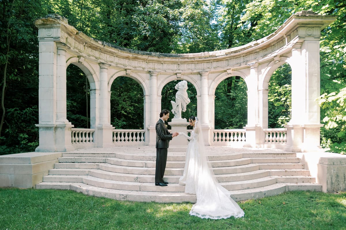 The bride in a long white veil and the groom in a dark suit face each other before a curved stone colonnade with a statue.