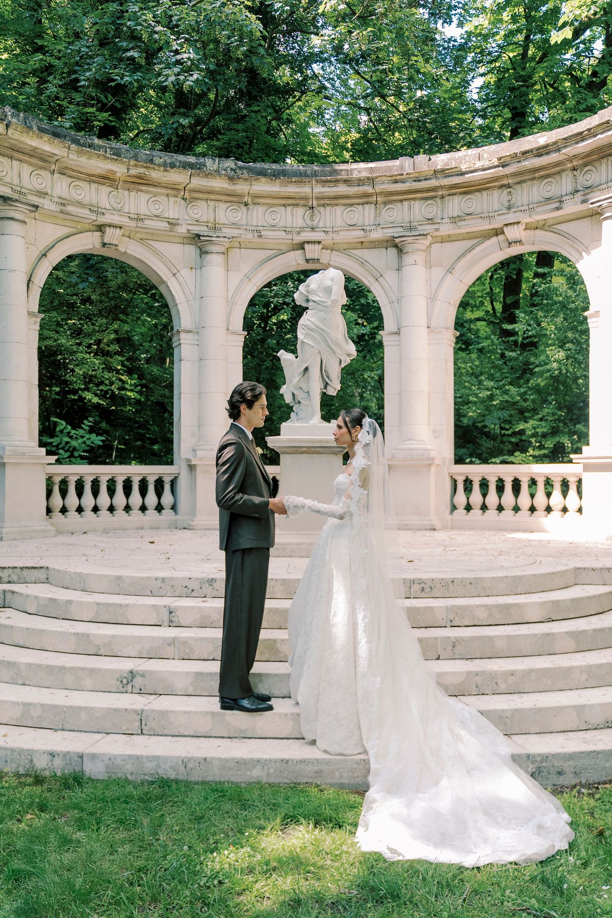 The couple stand facing each other on stone steps in front of a curved colonnade with a white statue between the arches.