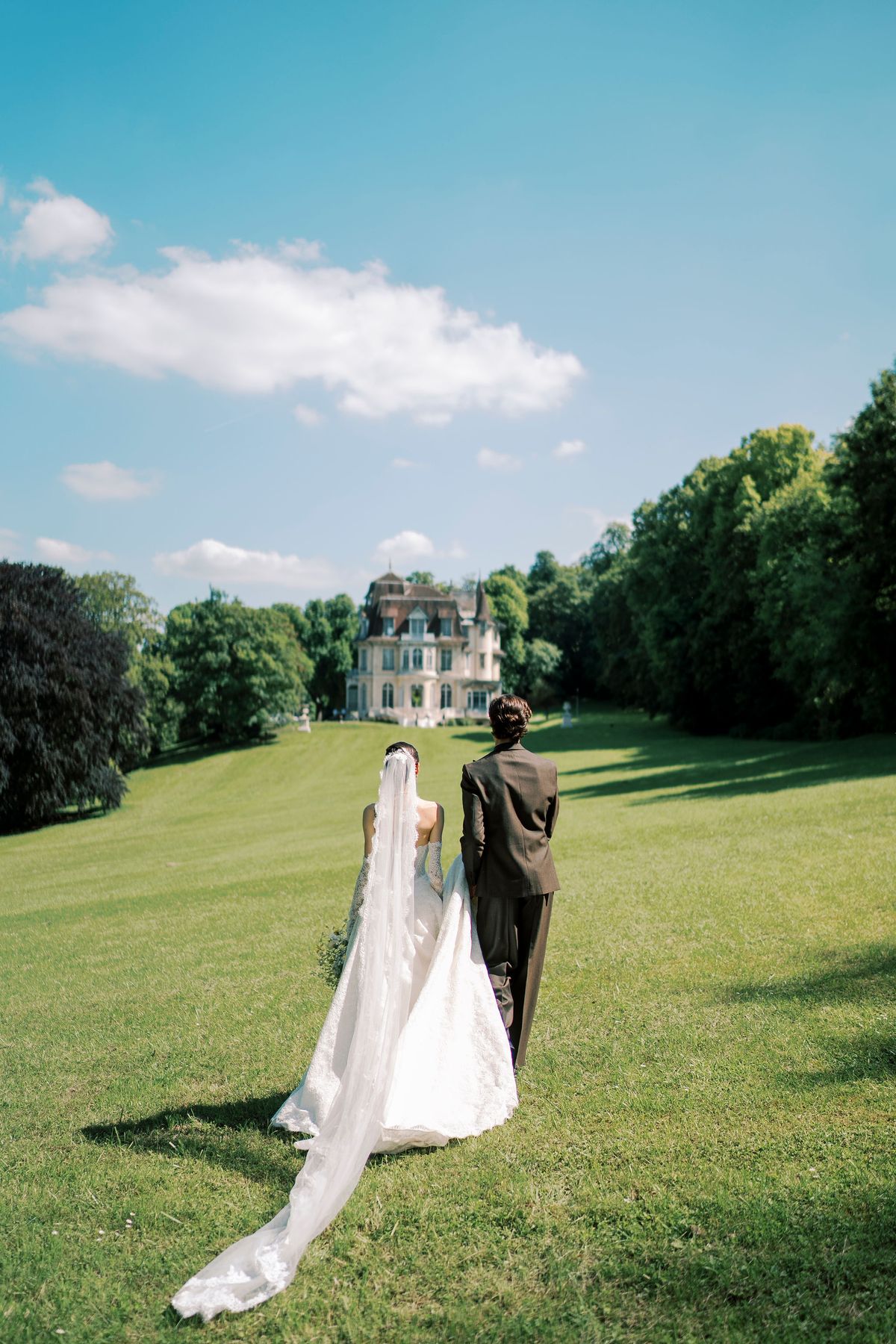 Seen from behind, the couple walk across a wide green lawn towards a distant chateau, the bride's veil trailing behind her.