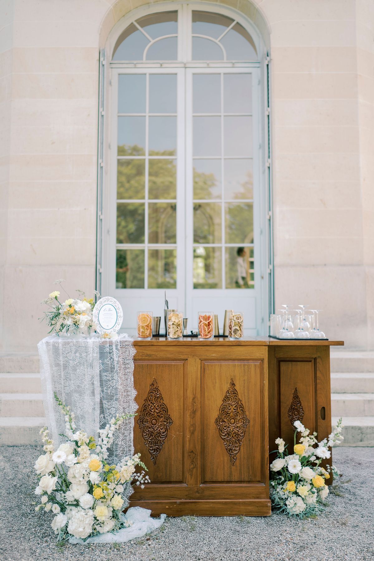 A carved wooden sideboard topped with lace, glassware and bottles stands beneath a tall arched window, flanked by white and yellow flowers.