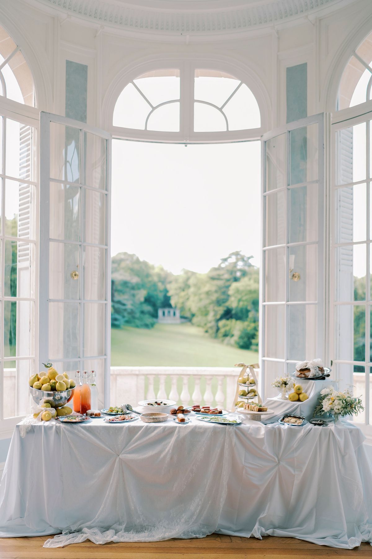 A long table with a grey cloth holds food displays and flowers beneath tall arched windows that look out over a green park.