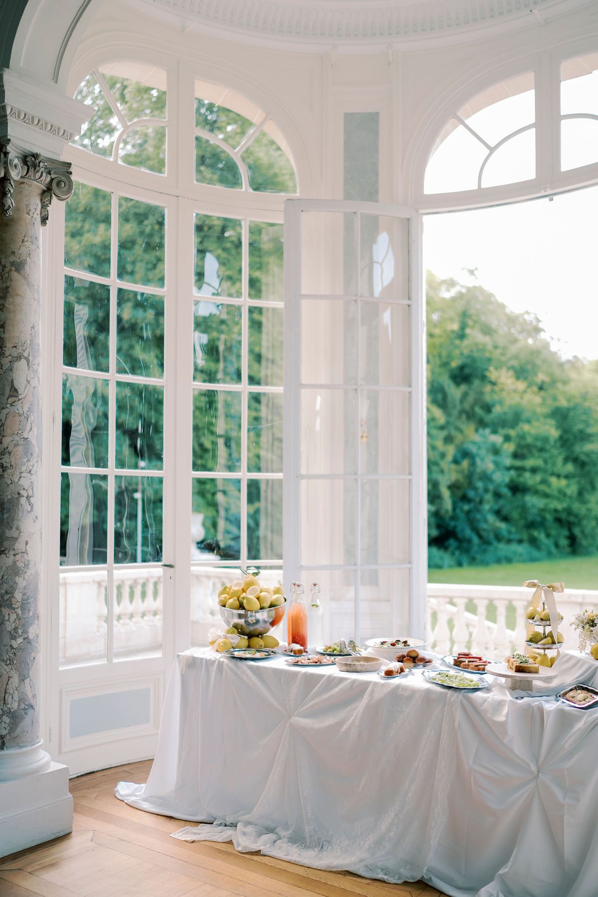 A table with a white cloth holds a menu, food and drinks beside tall windows that look out onto trees.