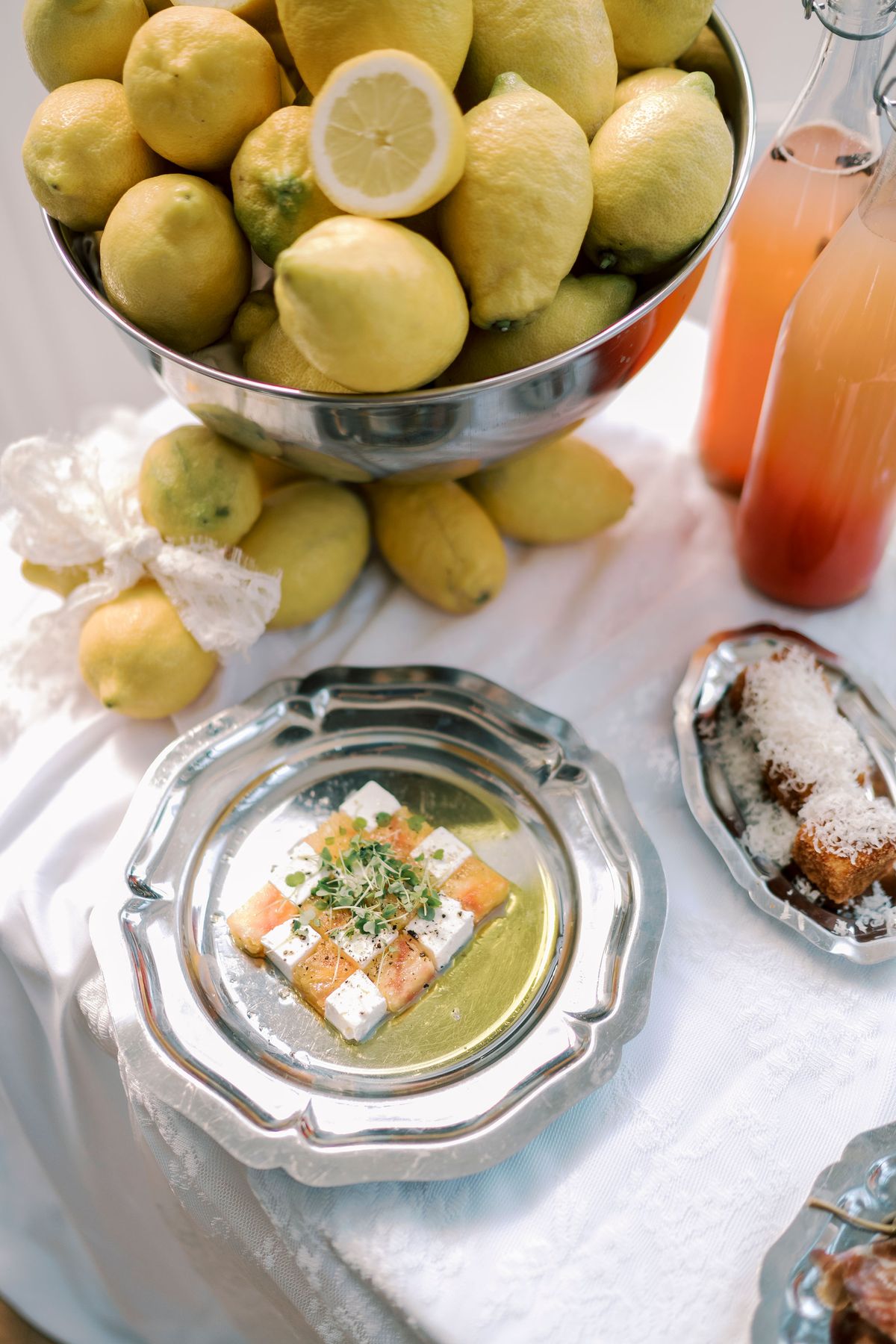 A steel bowl piled with lemons stands beside bottles of pink drink and pewter plates holding small savoury dishes on white linen.