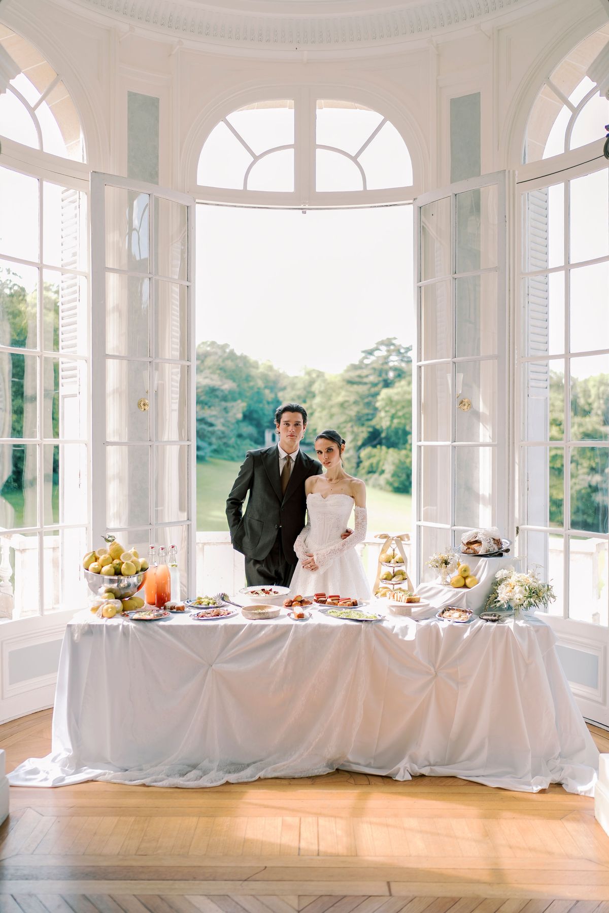 The couple stand in a bright window-lined rotunda behind a long white-draped table of food, with parkland visible outside.