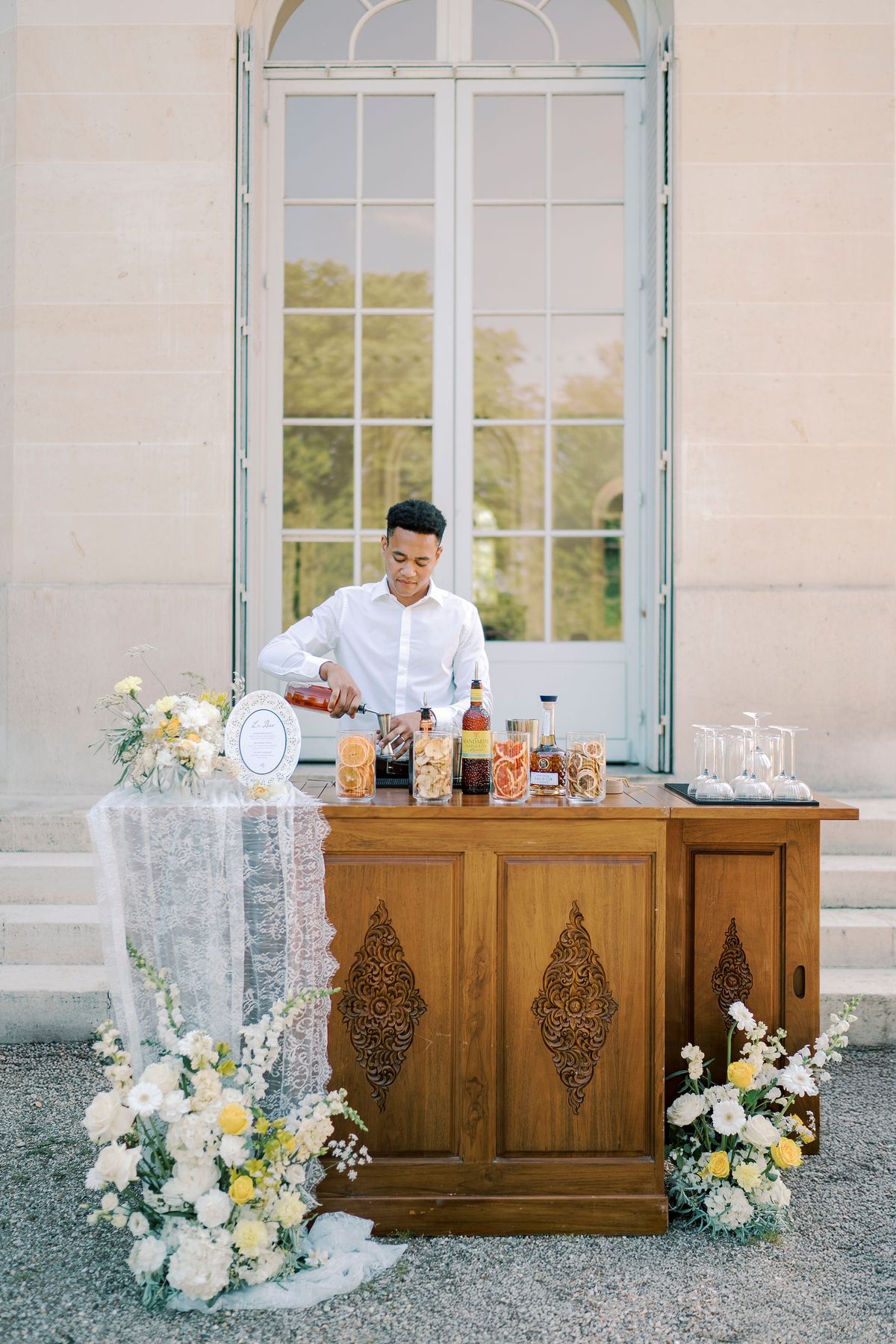 A bartender in a white shirt prepares drinks at a carved wooden bar decorated with white and yellow flowers and lace, beside tall windows.