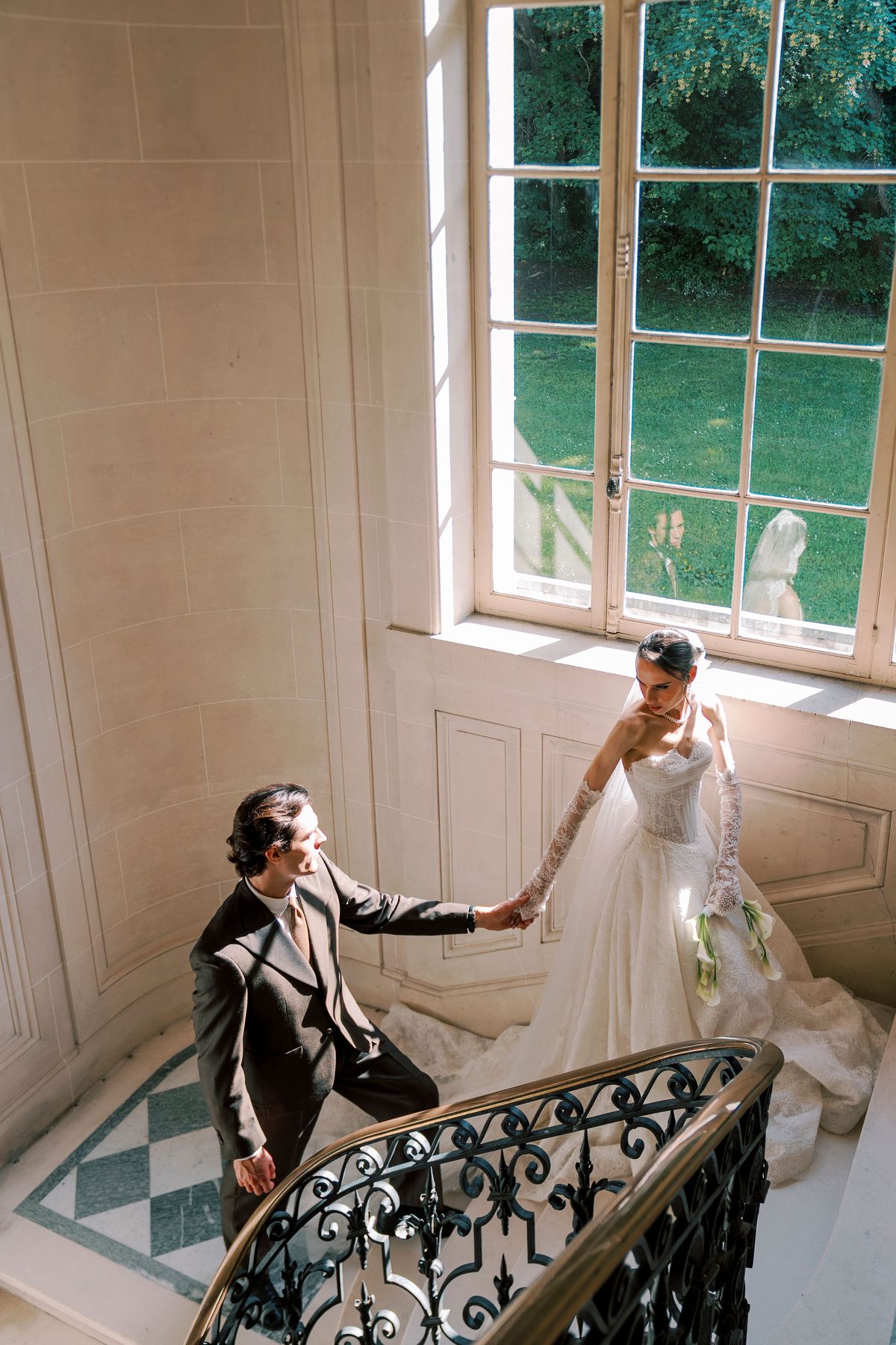 The groom sits on a stair landing holding the bride's hand as she stands beside him, near a tall window and an ornate iron banister.