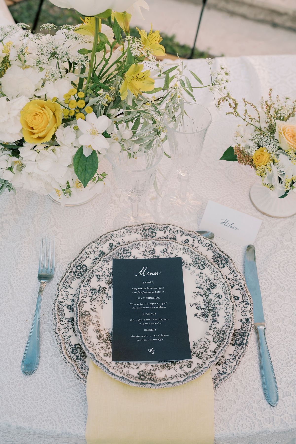 A table setting with a patterned blue and white plate, a dark menu card, light blue cutlery, a yellow napkin and yellow flowers.