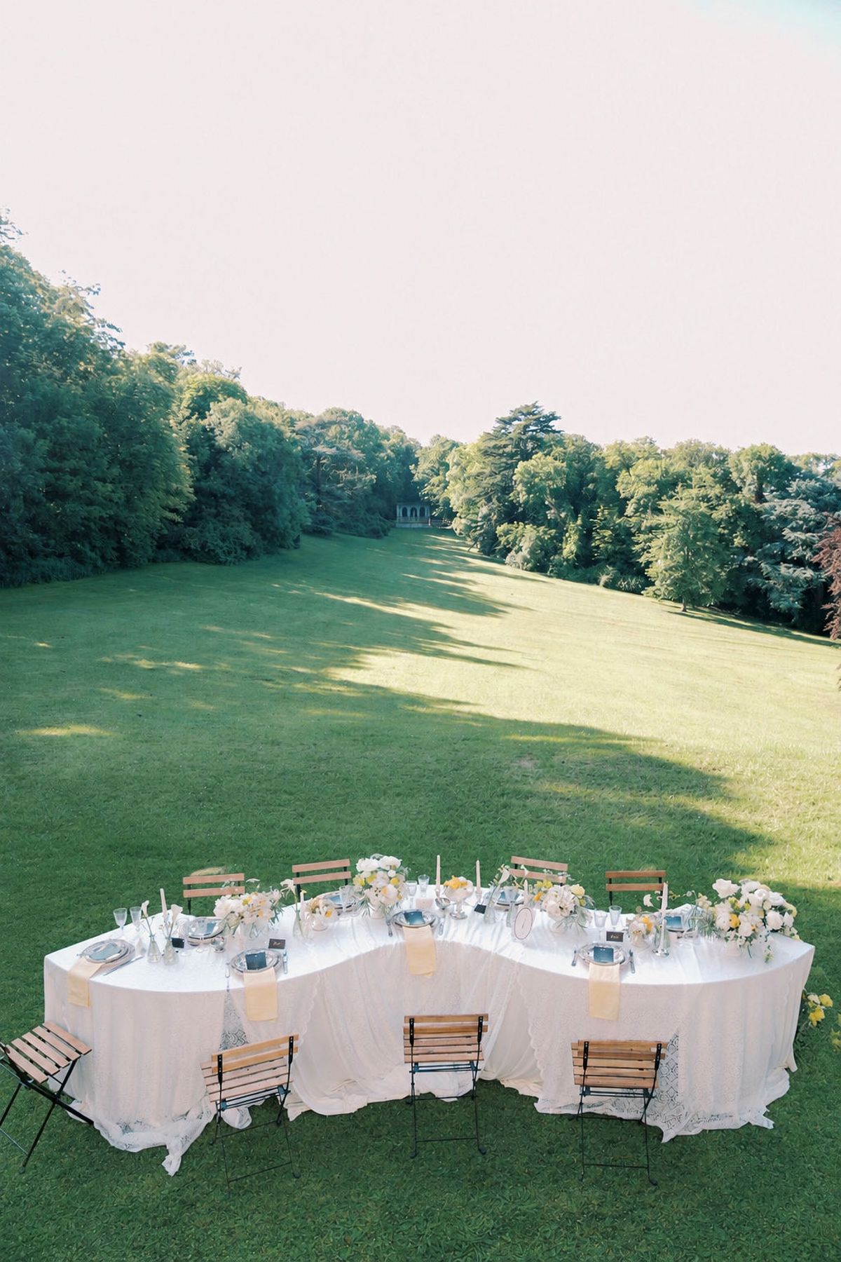 A long, curving table dressed in white linen with wooden chairs stands on a sunlit lawn bordered by tall trees.