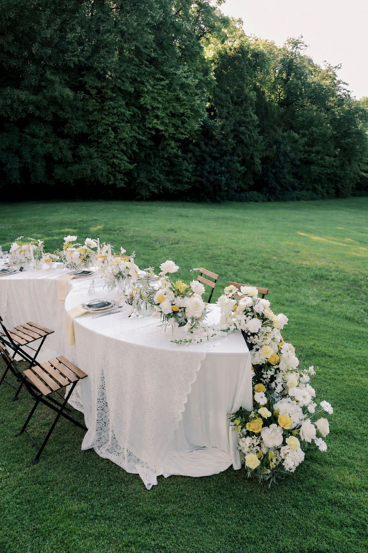 A curved table with white linen and lace is edged with a garland of white and yellow flowers on grass, with folding chairs nearby.