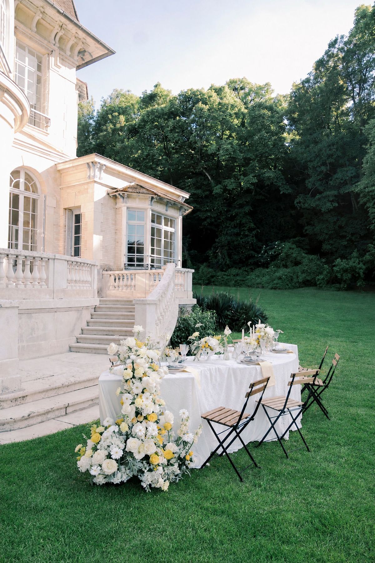 A round table with white linen, yellow and white flowers and folding chairs on a lawn beside a pale stone manor house.