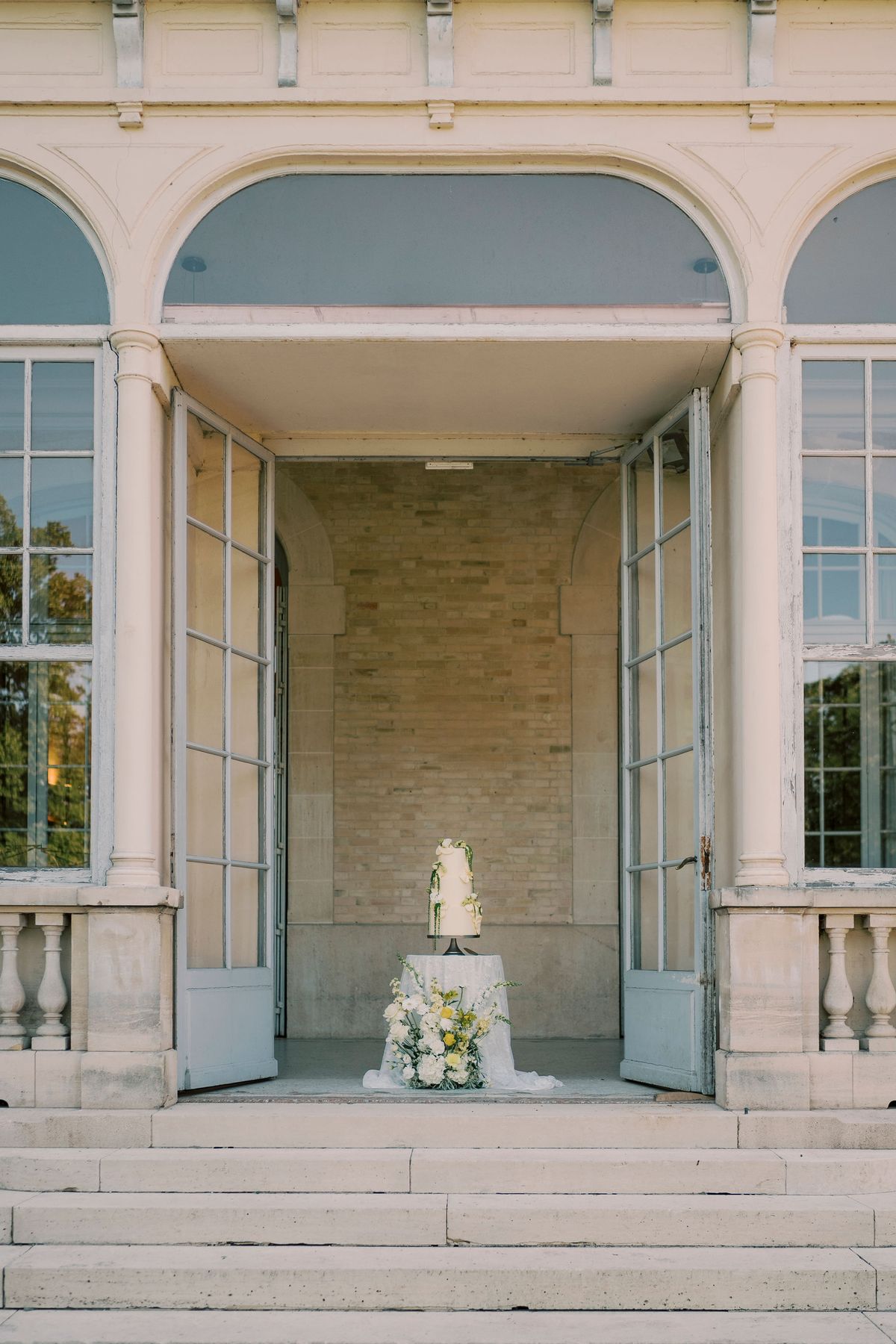 A tiered white cake on a small round table stands in the open doorway of a stone pavilion at the top of steps.