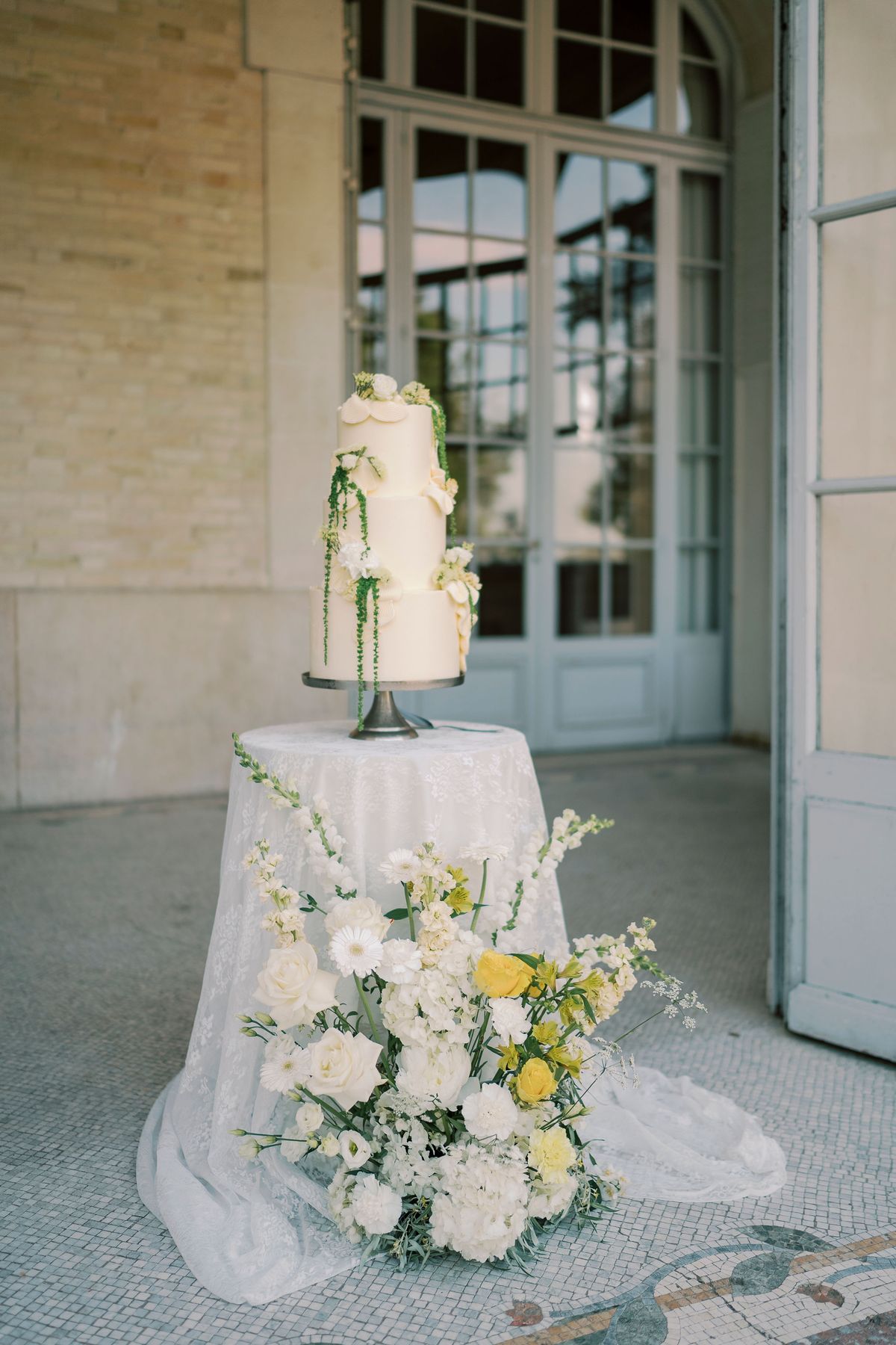 A white tiered cake sits on a linen-draped table with a large arrangement of white and yellow flowers, before tall glass doors.