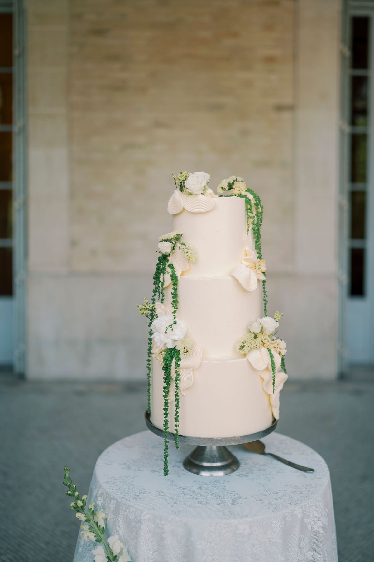 A tall white tiered cake decorated with cream flowers and trailing green stems stands on a lace-covered table against a pale stone wall.