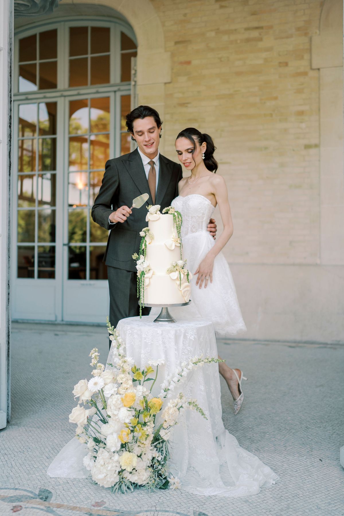 The couple stand together cutting a tall white cake in front of French windows, with yellow and white flowers in the foreground.