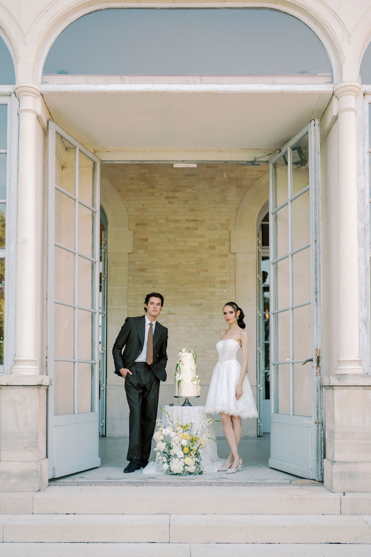 The couple stand side by side beside a tiered cake in the open doorway of a stone pavilion framed by columns.