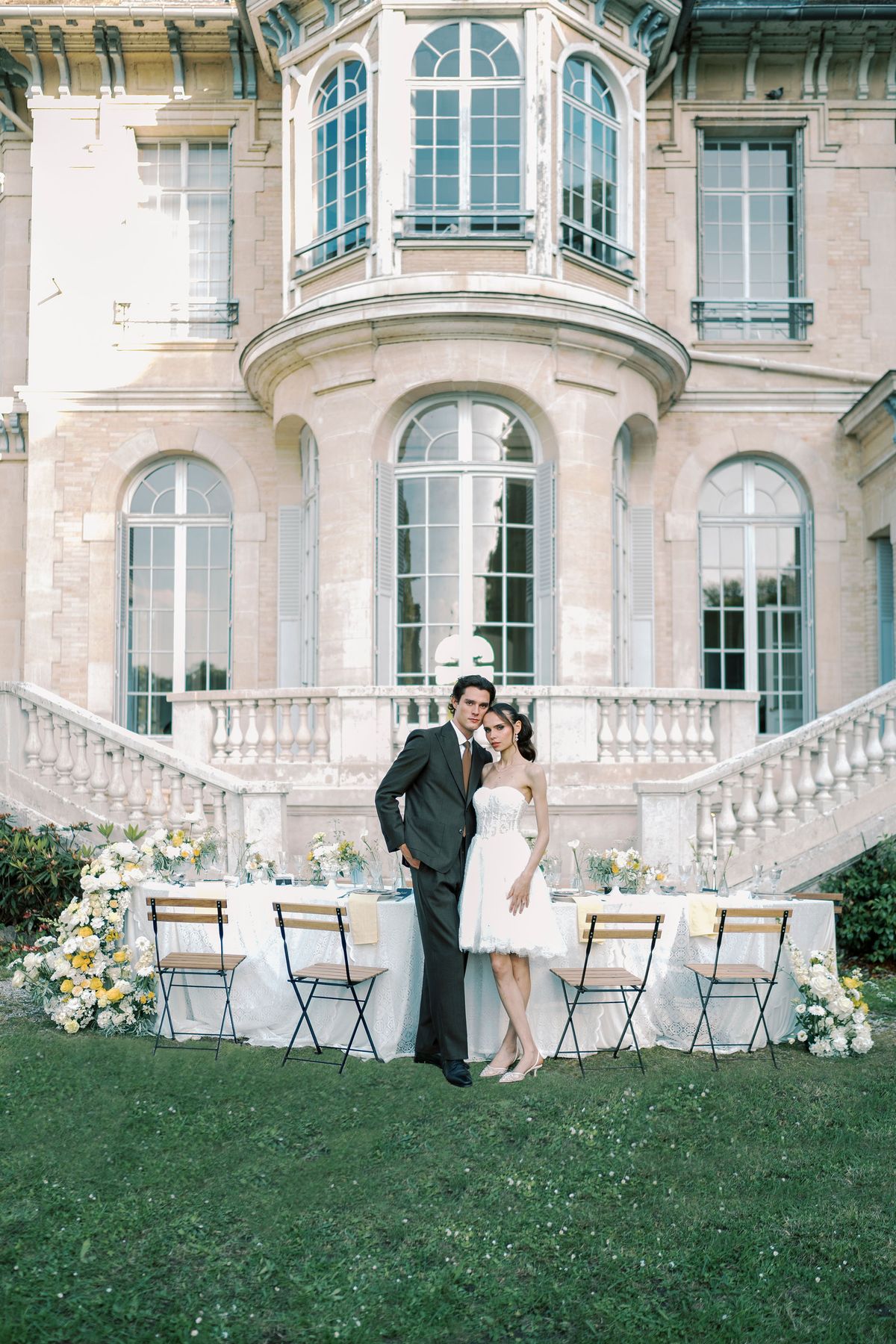 The couple stand together on the lawn in front of a pale stone chateau, beside a table dressed with white linen and yellow flowers.