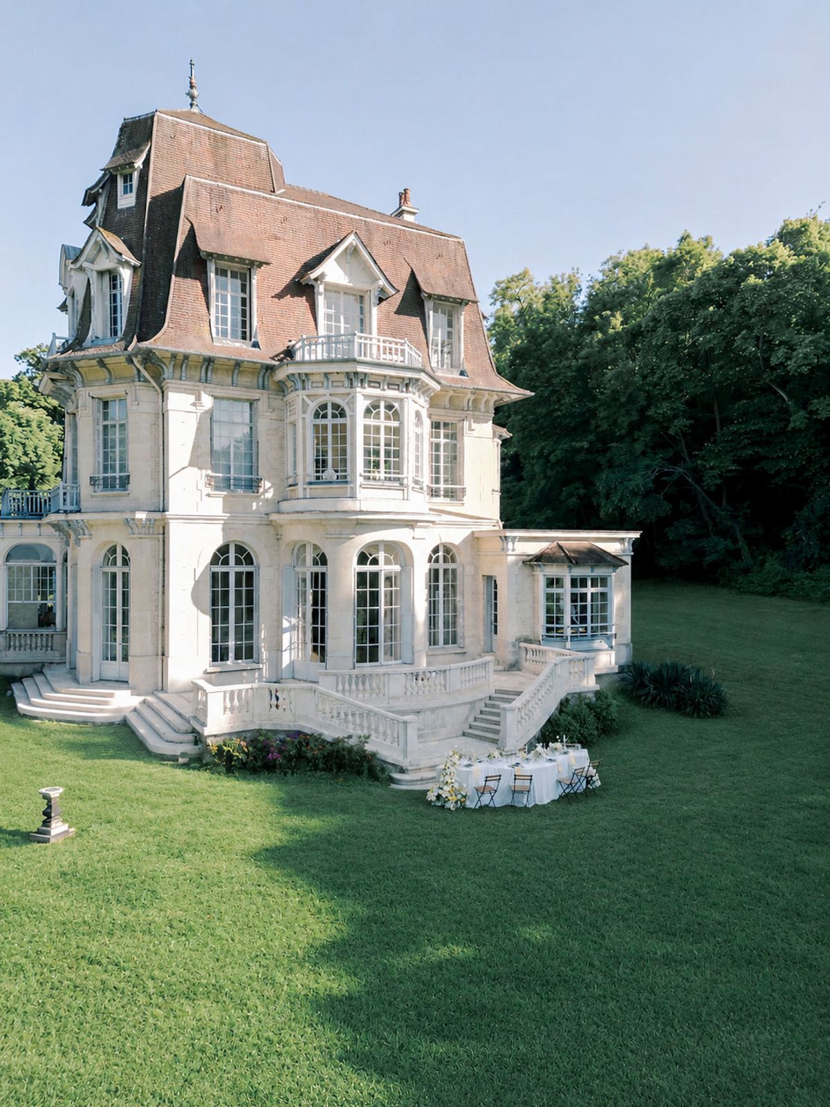 A cream stone chateau with a mansard roof and tall windows stands beside a green lawn and trees under a blue sky.