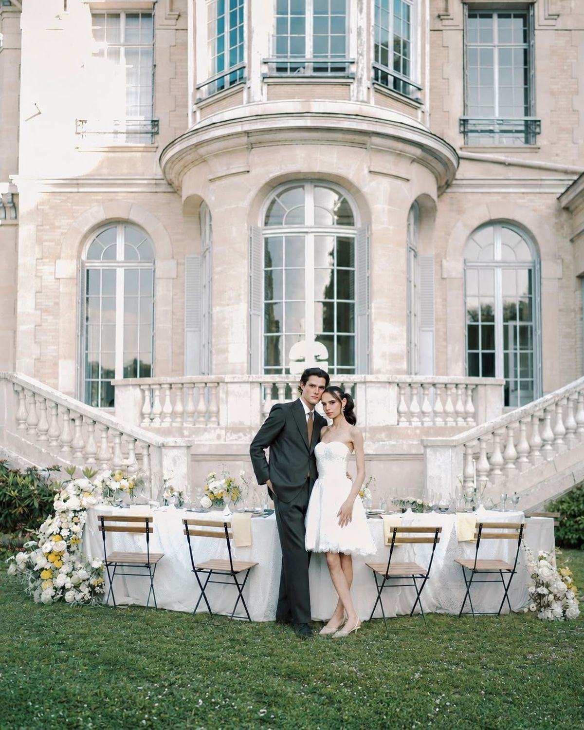 The couple stand side by side before a stone chateau with a curved facade, next to a table with folding chairs and flowers.