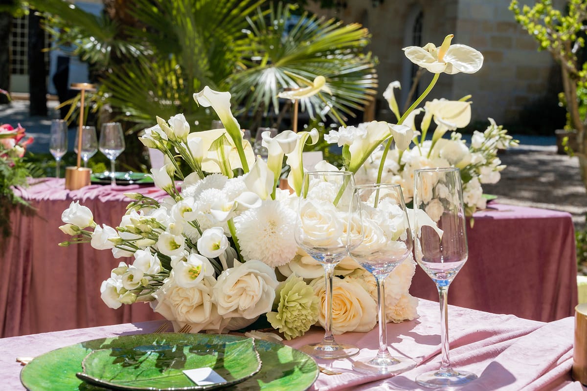 White floral centrepiece with roses and calla lilies beside coupe glasses on mauve velvet