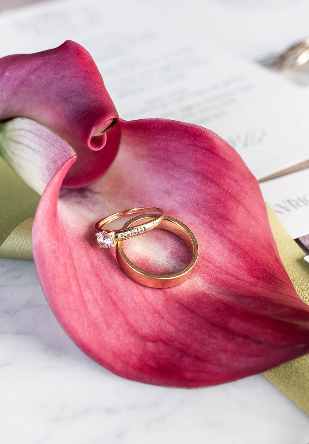 Wedding rings resting on a raspberry calla lily petal beside the stationery suite
