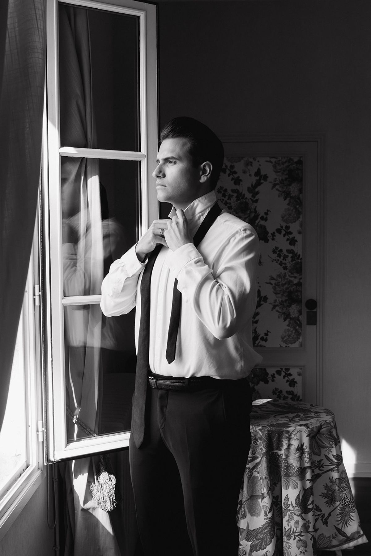 Black and white portrait of the groom fastening his tie at a window